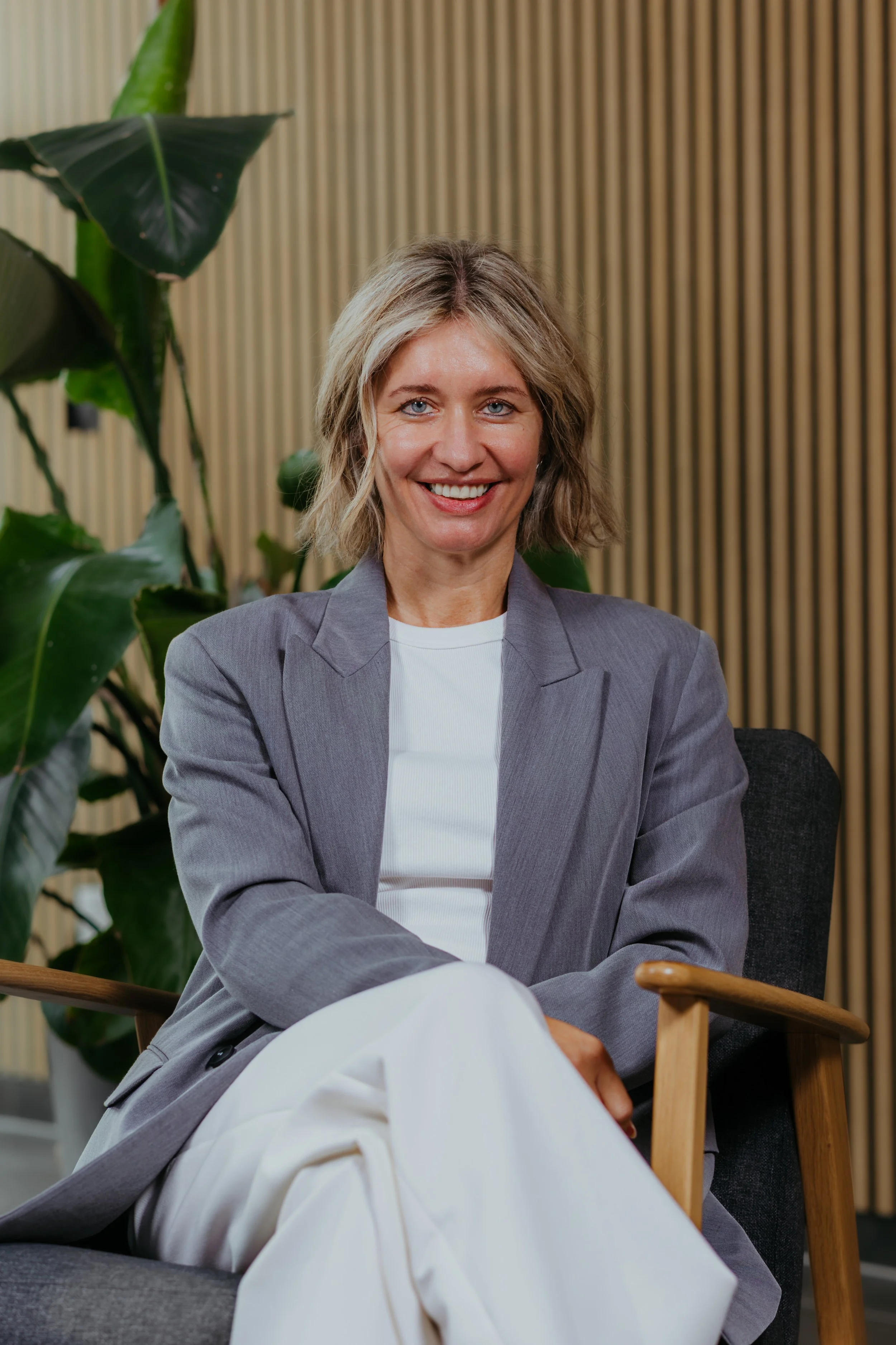 Vrouw zit in een stoel, glimlacht, draagt een grijze blazer en witte broek, met grote groene planten op de achtergrond en een houten wand.