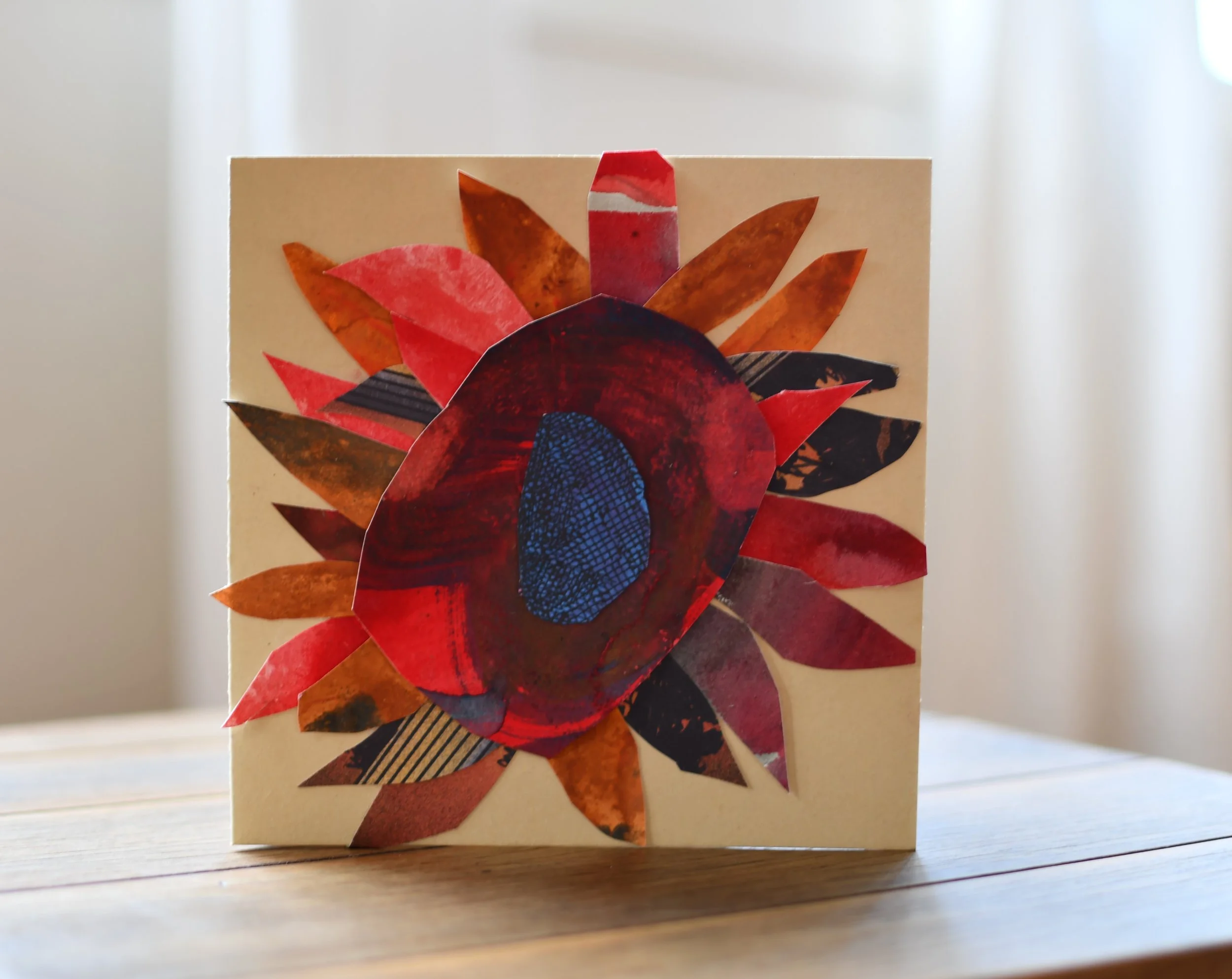 A handmade collage art piece of a flower with multi-colored paper petals on a beige background, displayed on a wooden surface.