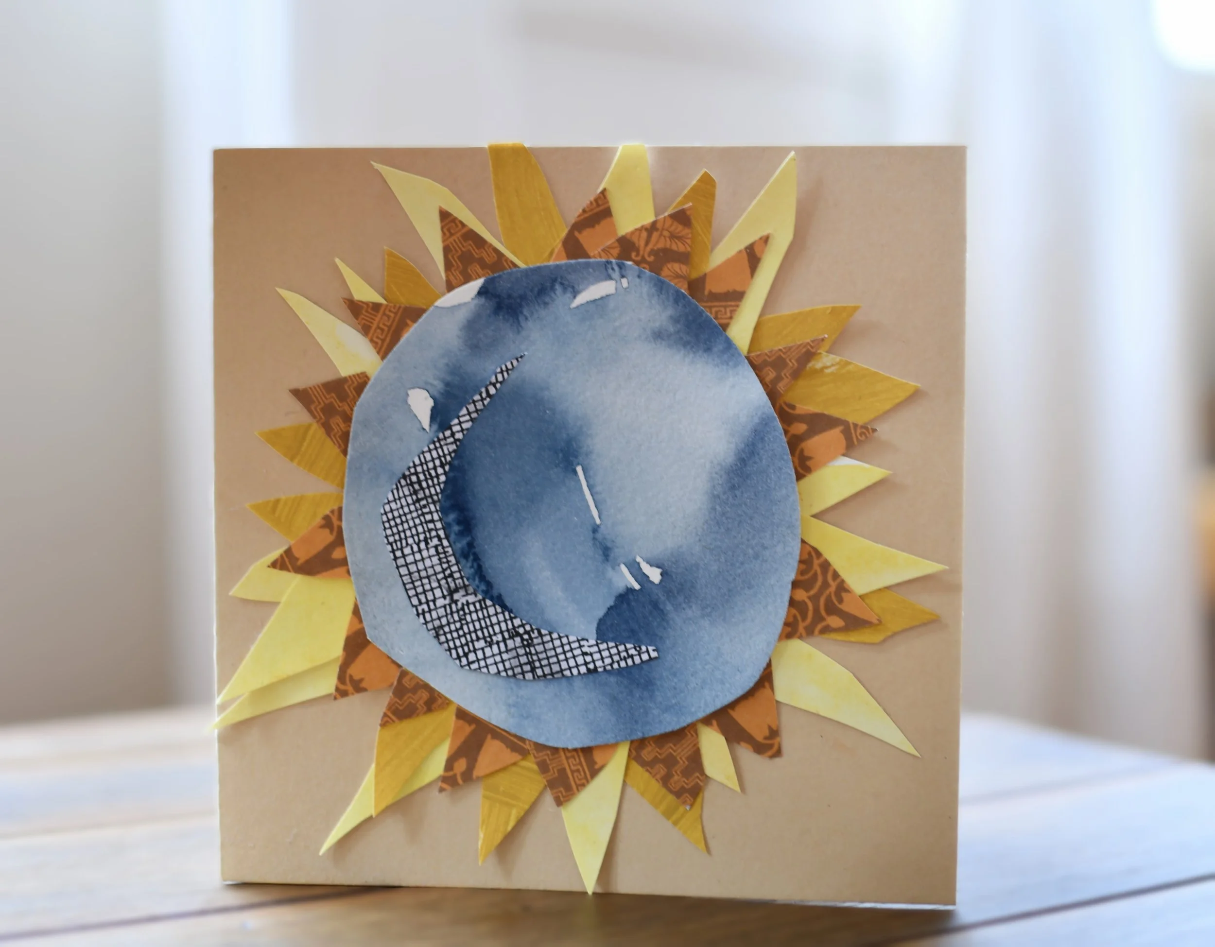 A handmade card with a sunflower design, featuring a blue watercolor moon with a smiling face and a black-and-white checkered crescent moon pattern, surrounded by yellow and brown paper petals.