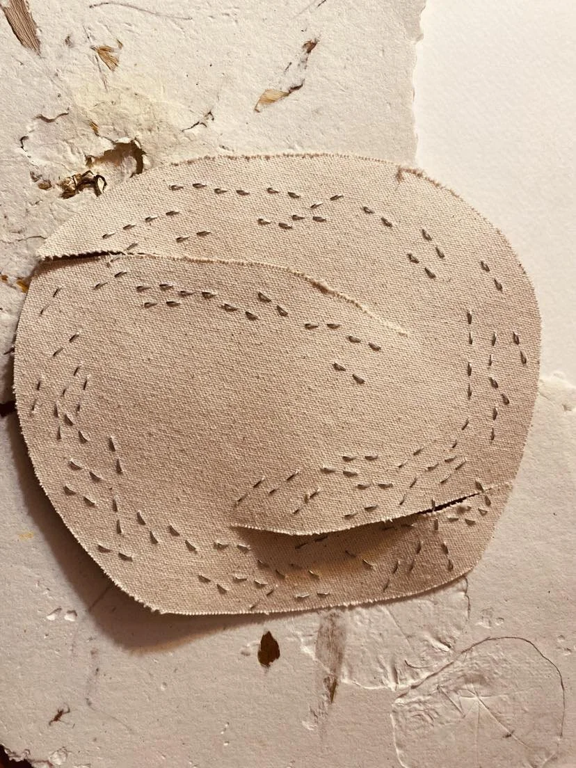 A beige, textured fabric patch with evenly spaced cut-out stitching, placed on a distressed, white, painted wall surface.