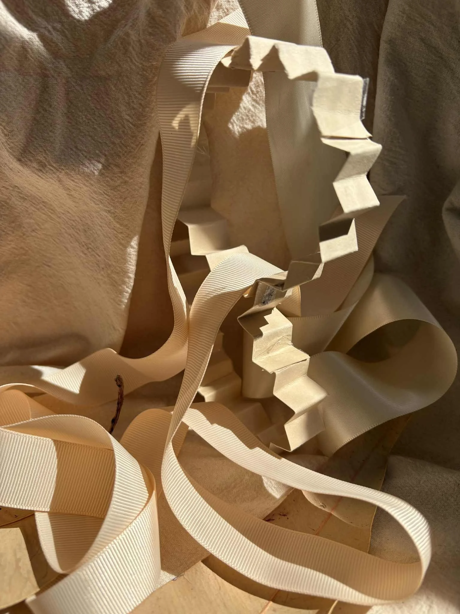 A cream-colored ribbon and a white foam trimming are tangled and lying on a textured beige surface, with some parts of the ribbon curled and some cut into zigzag shapes.