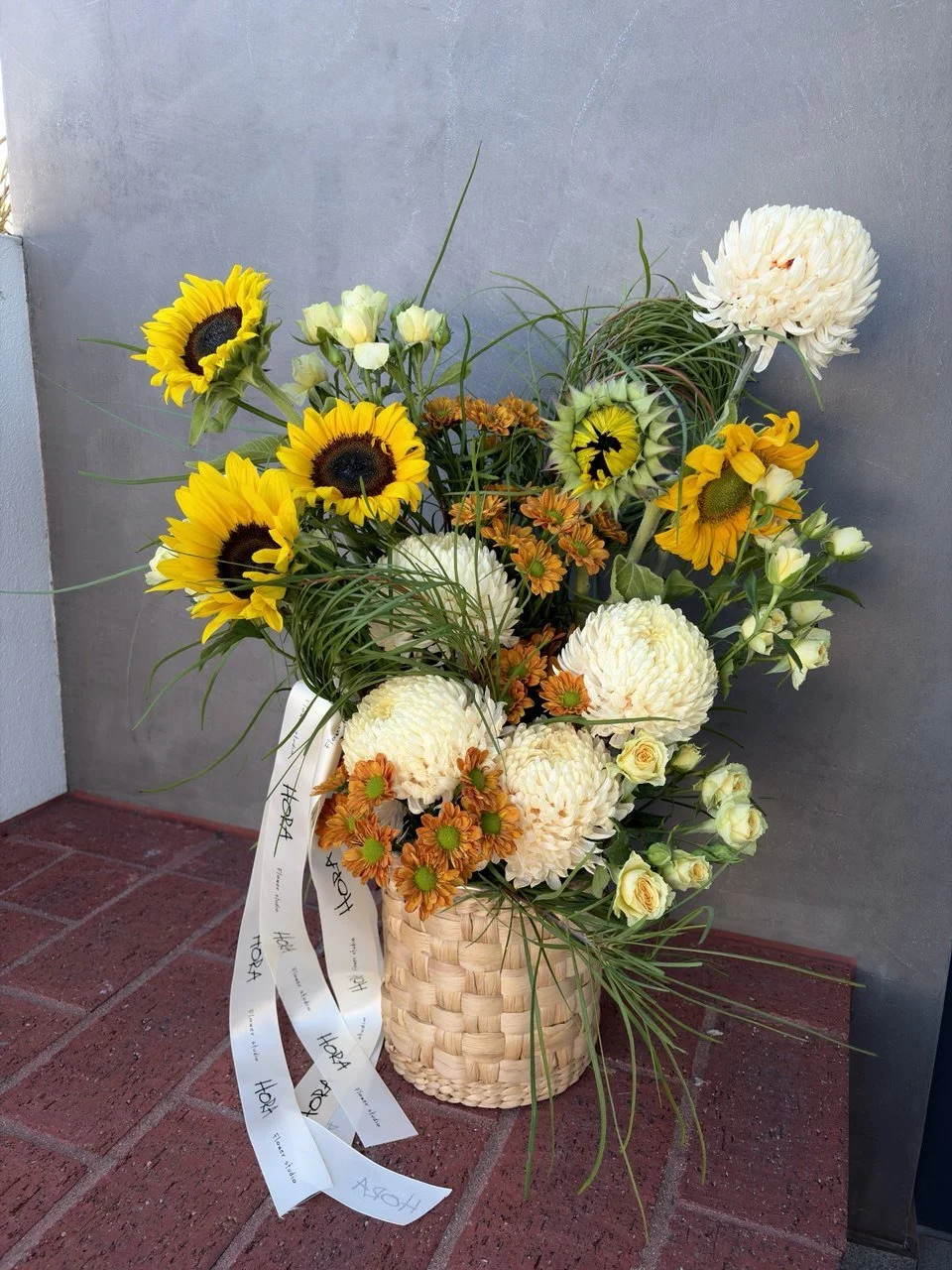 A seasonal sunflower basket arrangement by HORA Flowers in Perth, held in front of a brick wall with branded ribbon detail.