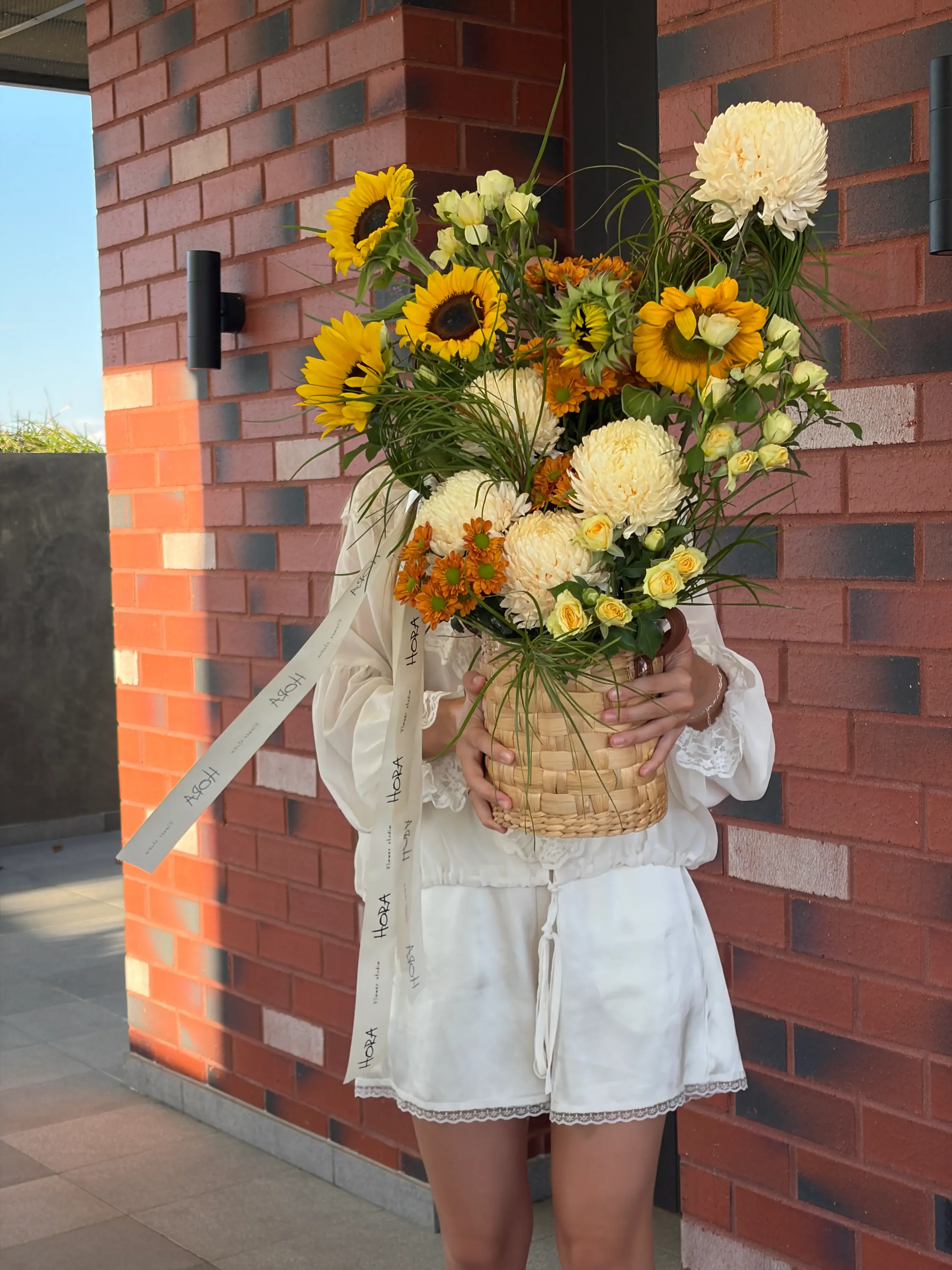 Seasonal sunflower basket arrangement from HORA Flowers in Perth, styled on brick with a soft grey wall backdrop.