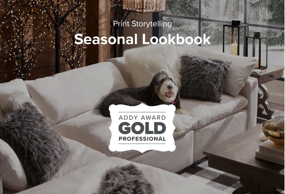 Cozy living room with white sofa, dark cushions, white furry pillows, a happy dog laying on the sofa, wooden coffee table with decorative items, and floor-to-ceiling windows showing a snowy landscape outside.