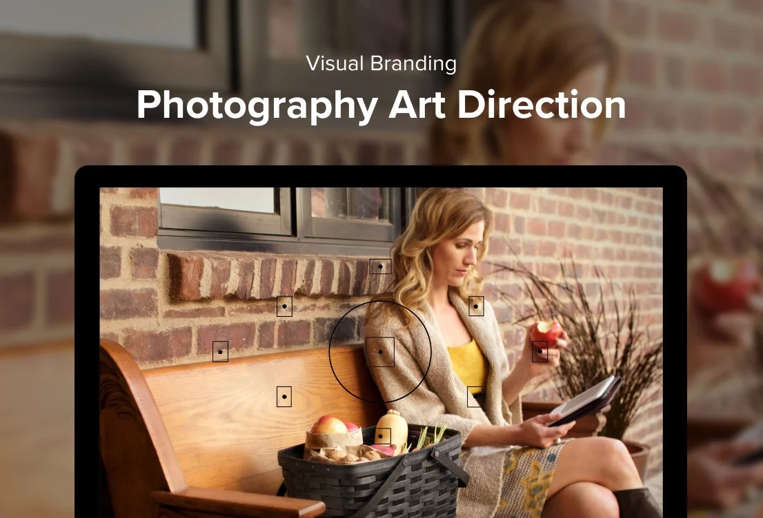 A woman sitting on a wooden bench outside a brick building, using a tablet device, with a basket of apples and other items beside her.