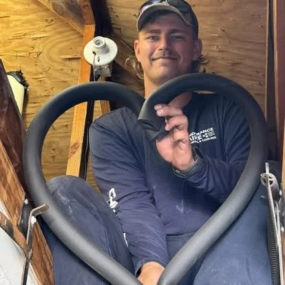 A young man sitting inside a wooden shed, holding a large black bow, dressed in outdoor gear including a cap and jacket.