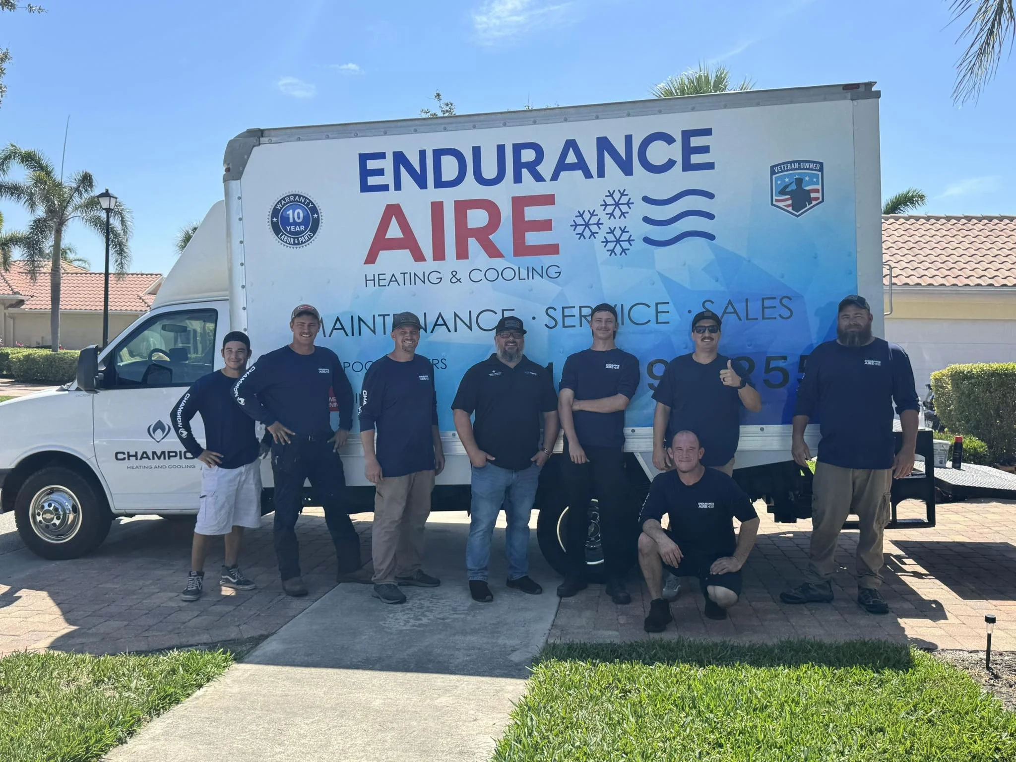 Group of seven men standing in front of a white service truck with blue and red signage for Endurance Air Heating & Cooling, outdoors on a sunny day with palm trees and houses in the background.
