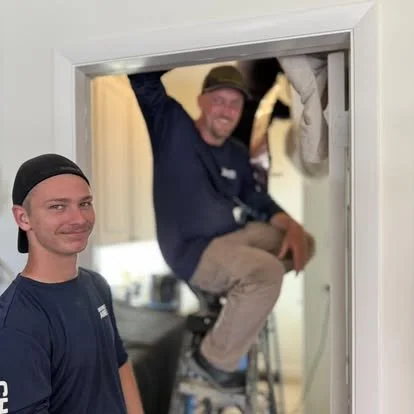 Two men working on a home renovation, one standing outside the doorway and the other sitting on a ladder inside, both smiling.