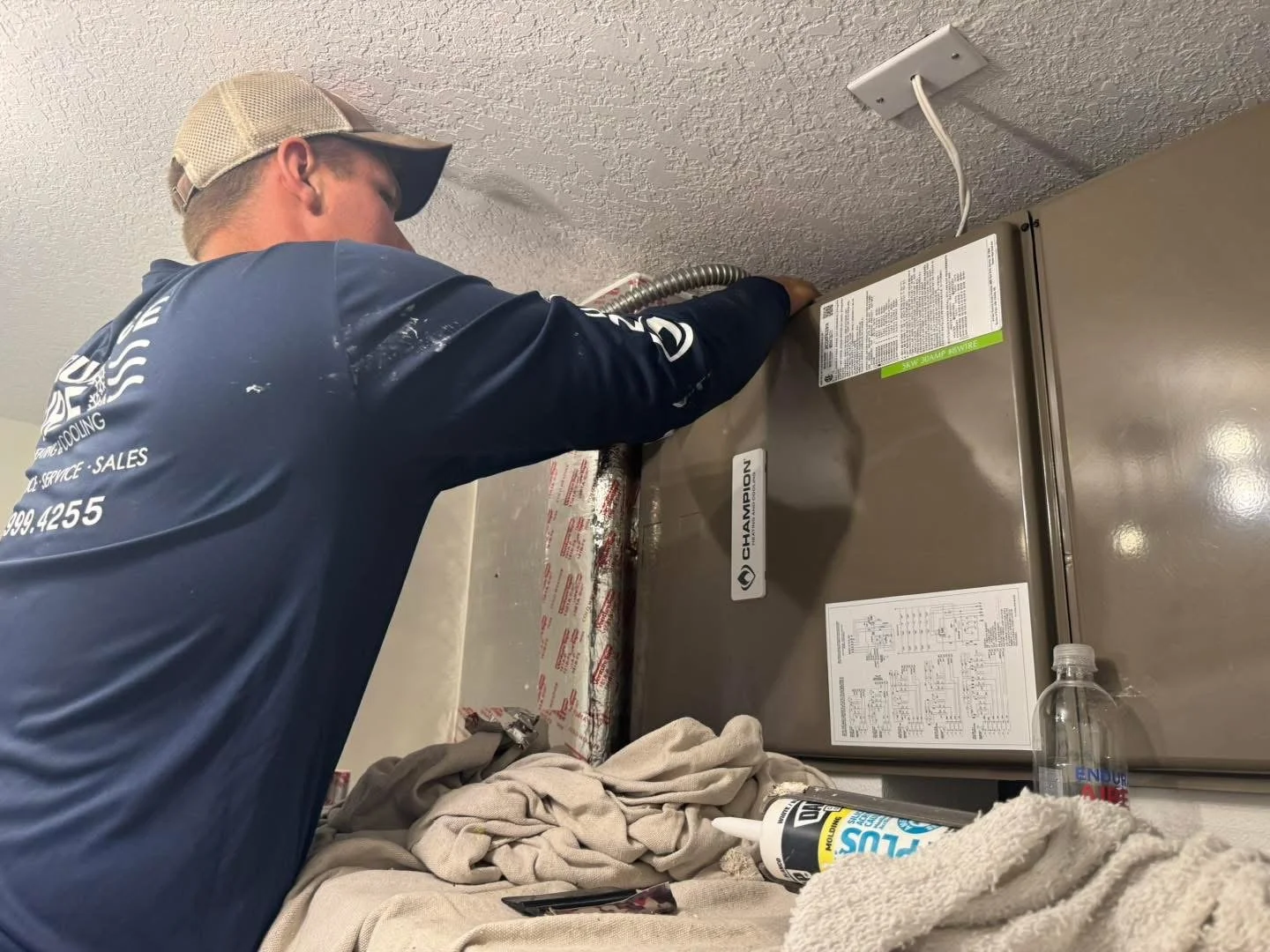 A technician in a navy blue shirt and beige cap installs or repairs an HVAC unit on a beige wall. Tools and supplies are on a cloth-covered surface below.