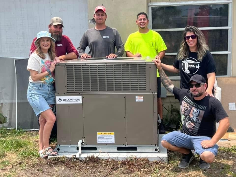 Group of six people standing and squatting around a large HVAC unit outside a house, smiling for the photo after they received a free ac system
