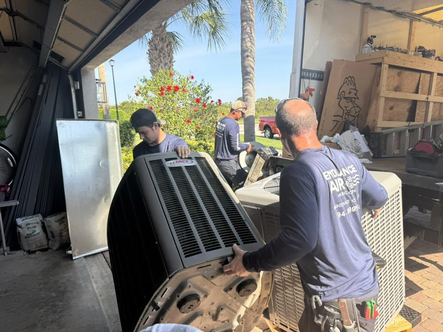 Three HVAC technicians working to install or repair air conditioning units outside a building, with palm trees and a flower bush in the background.
