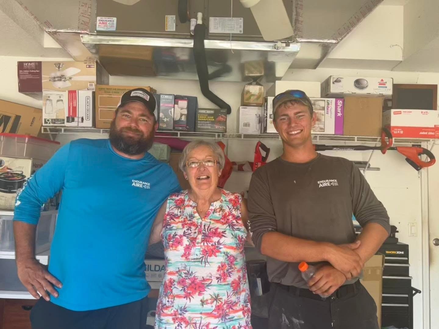 Three people, two men and one woman, smiling and standing together in a garage or workshop with shelves of boxes and tools behind them.