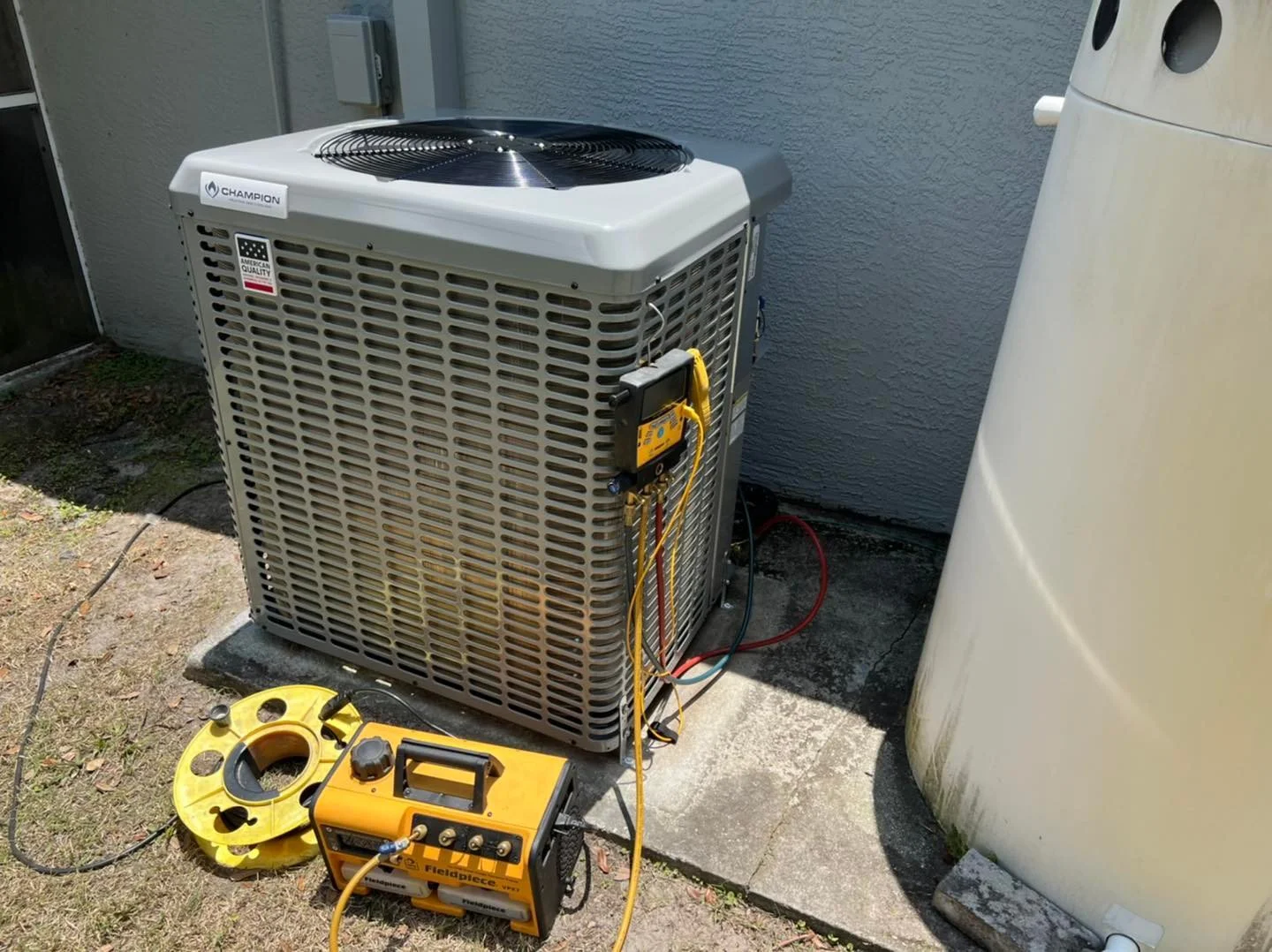 A central air conditioning unit outdoors next to a wall with connected gauges and tools for testing or maintenance.