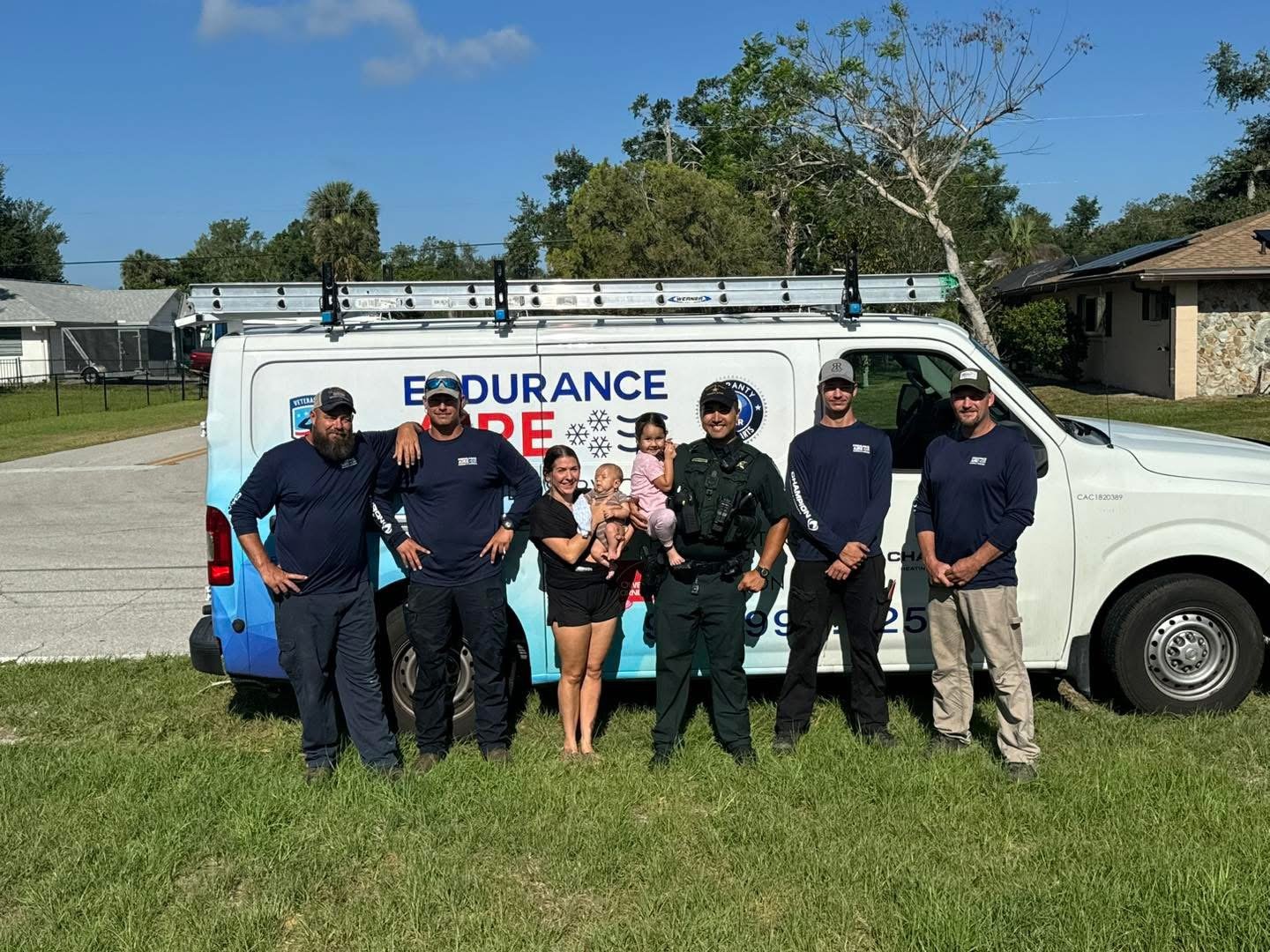 Group of six people, including children and a police officer, posing in front of an enduro fire safety service van on a sunny day, with houses and trees in the background.