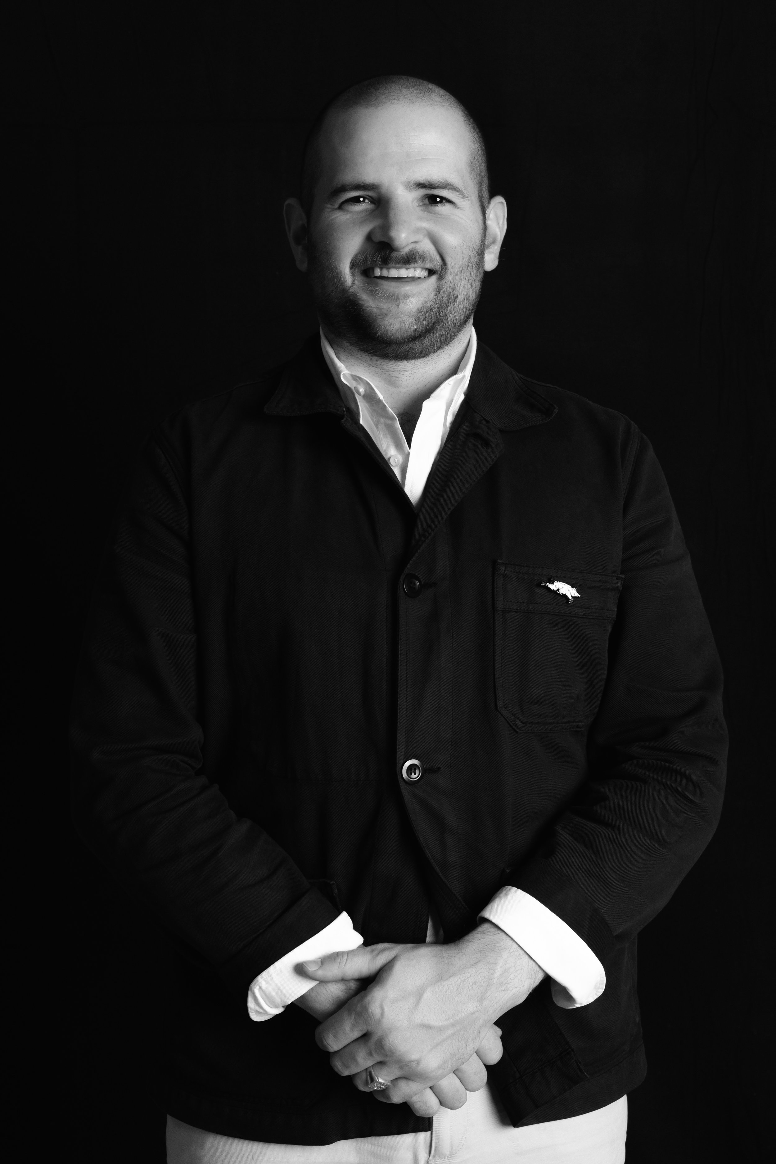 Schwarz-weiß Portrait eines Mannes mit kurzem Haar und Bart, trägt eine dunkle Jacke und weißes Hemd, lächelt. Hintergrund schwarz.