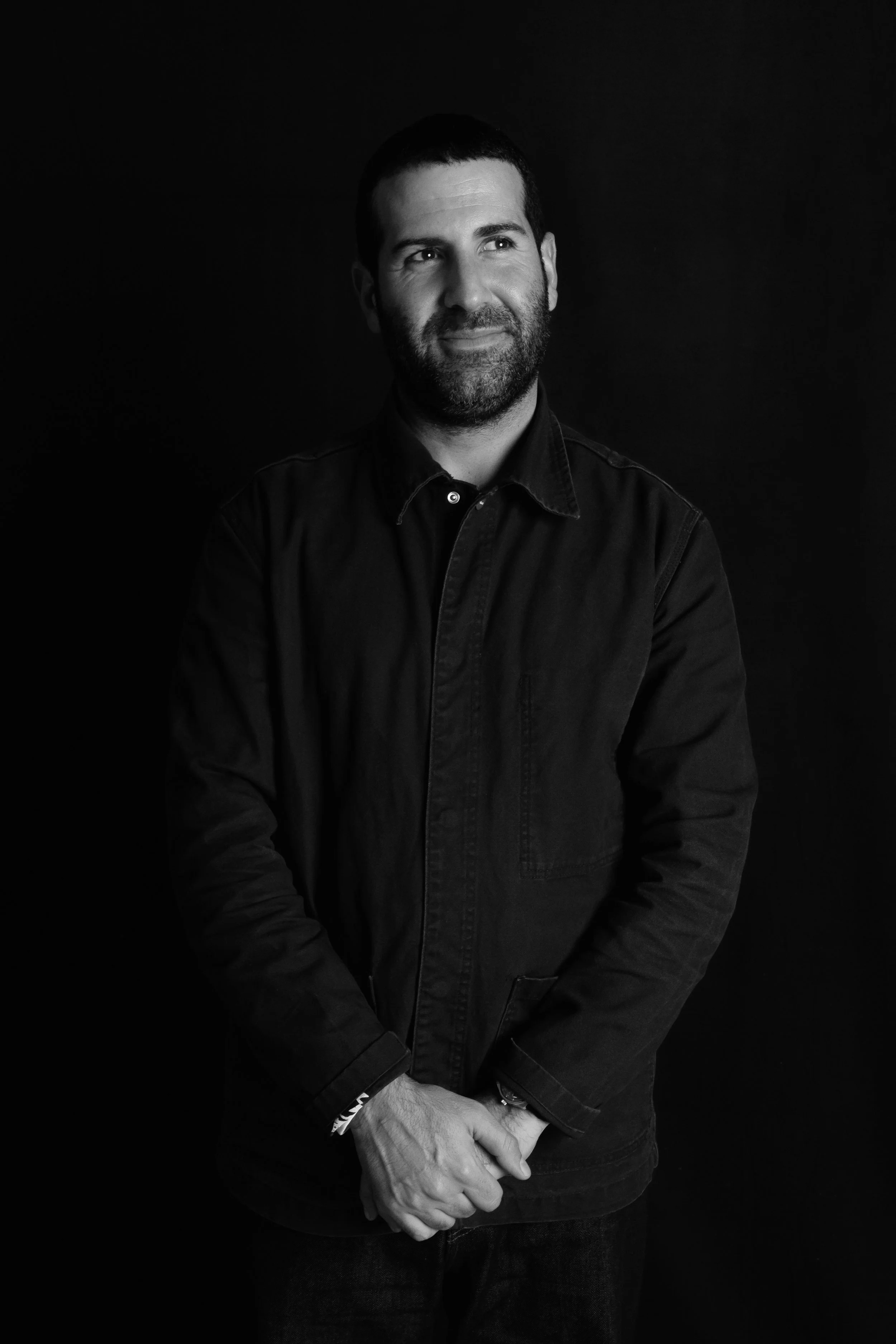 Schwarz-weiß-Porträt eines Mannes mit dunklem Hemd vor schwarzem Hintergrund, lächelt leicht, Hände vor Bauch verschränkt.