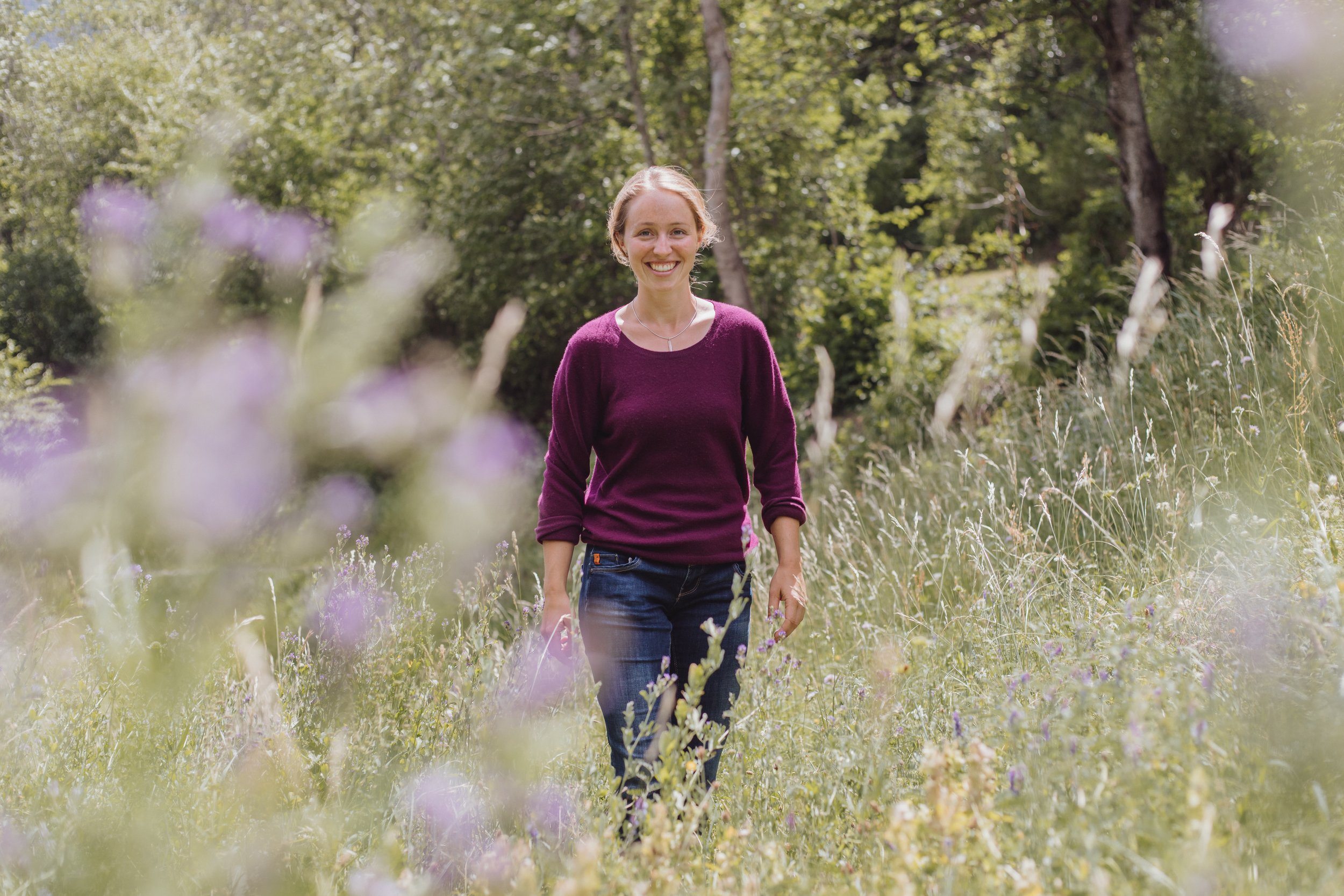 Junge Frau mit lila Pullover und Jeans, lächelnd, in einer grünen Wiese mit Blumen und Bäumen im Hintergrund.