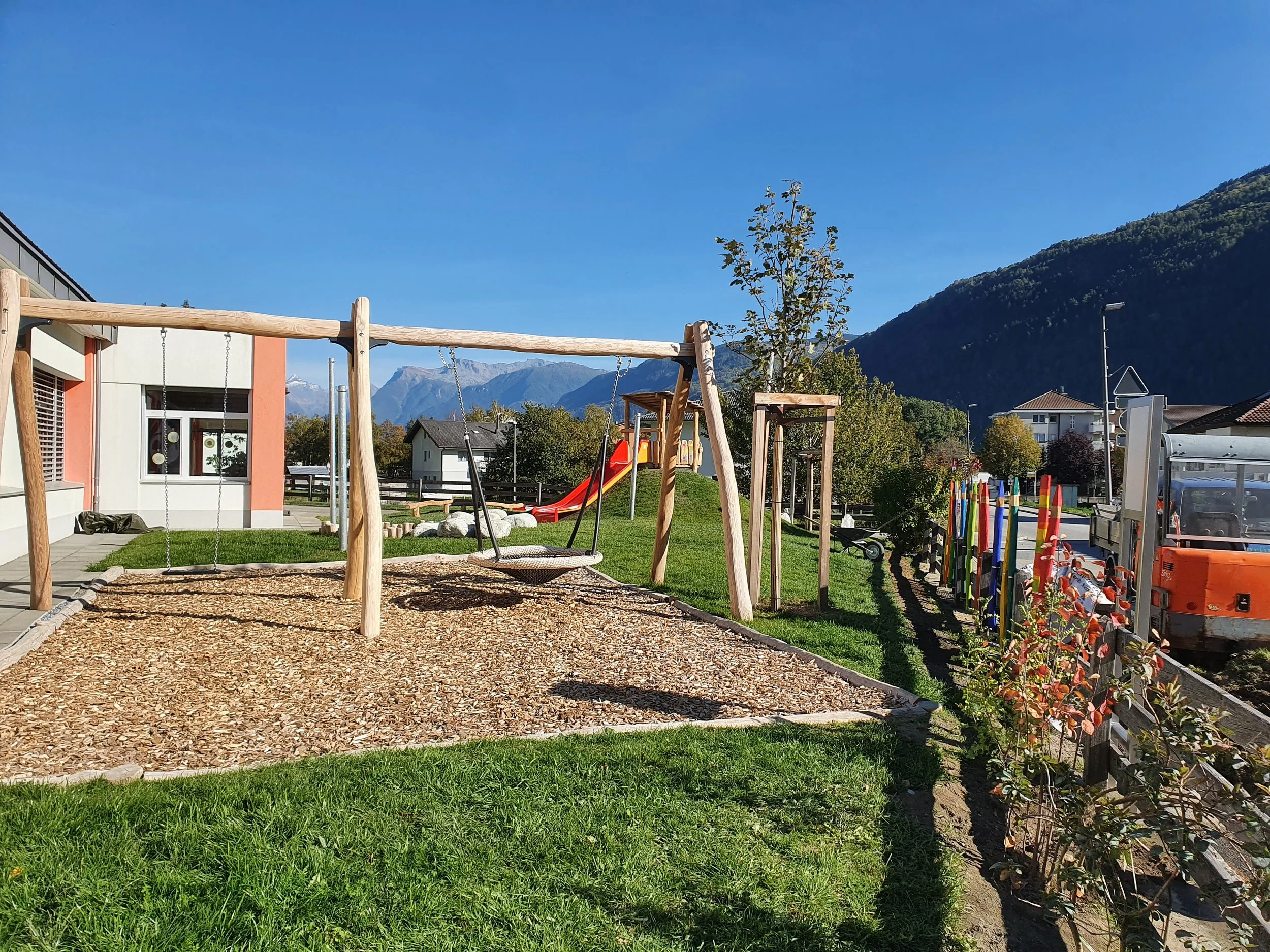 Ein Spielplatz mit Schaukeln, Rutsche und Klettergerüst im Freien, umgeben von Bäumen und einem Zaun, mit Bergen im Hintergrund an einem sonnigen Tag.