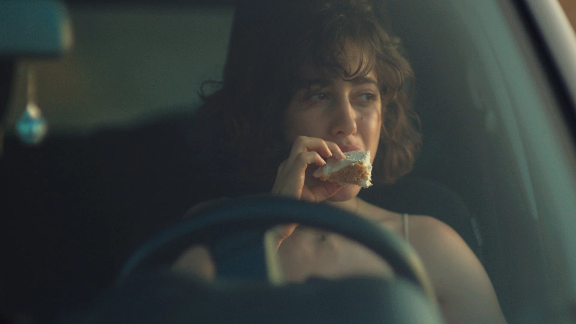 Femme avec cheveux bouclés mangeant un gâteau derrière le volant d'une voiture.