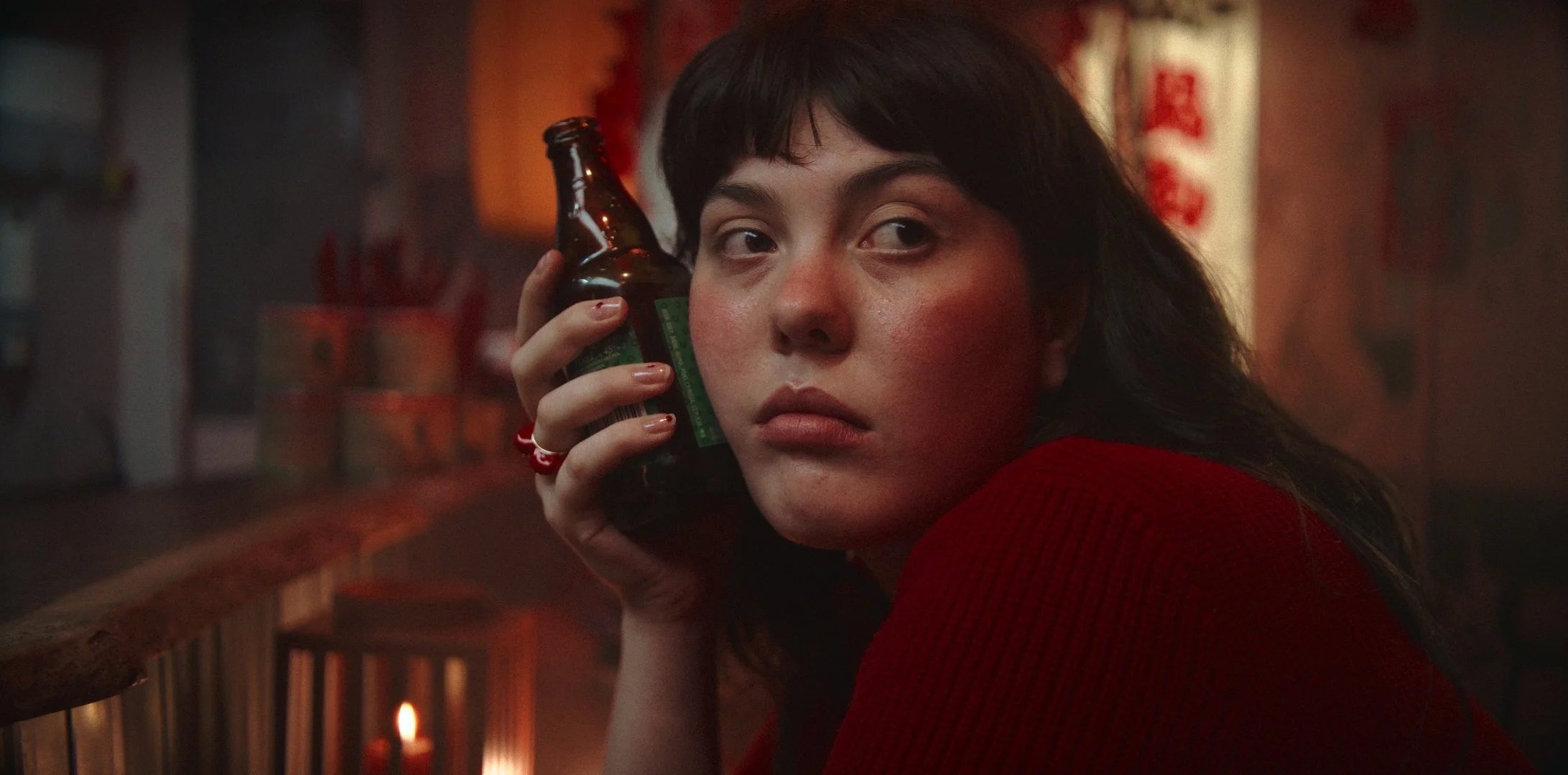 Une femme aux cheveux longs et noirs, portant un vêtement rouge, tient une bouteille de bière derrière son oreille dans un environnement sombre et chaud, avec des décorations de style asiatique en arrière-plan.