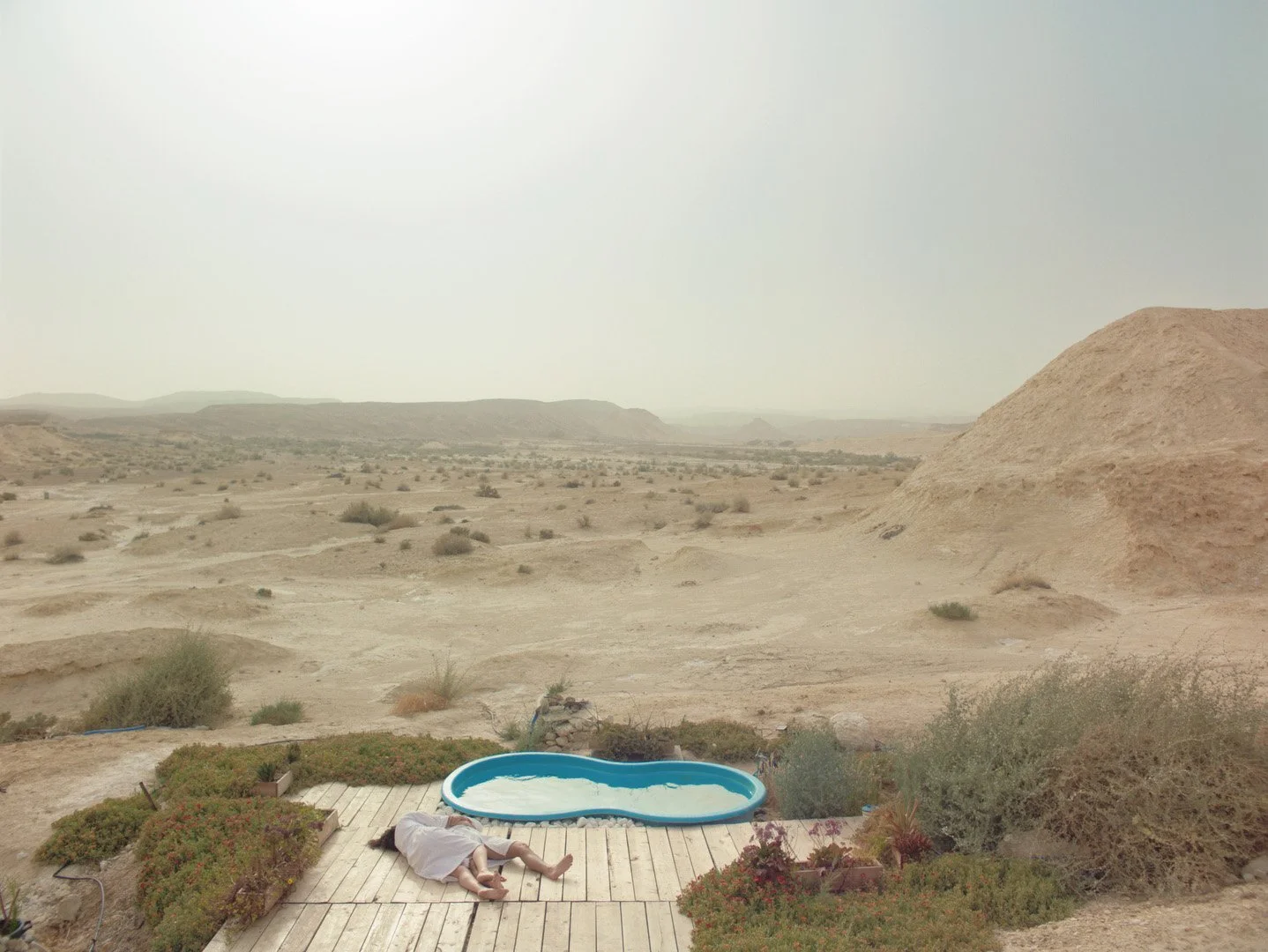 Une personne allongée sur un terrasse en bois près d'une petite piscine dans un désert avec des dunes et peu de végétation.