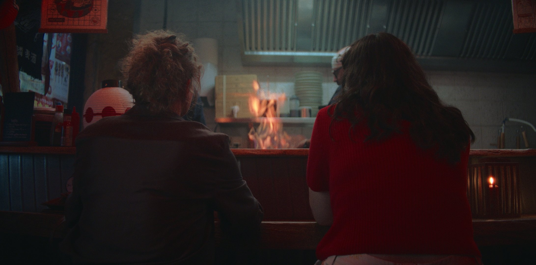 Deux personnes assises au comptoir d'un restaurant japonais, regardant une flamme dans la cuisine en arrière-plan.