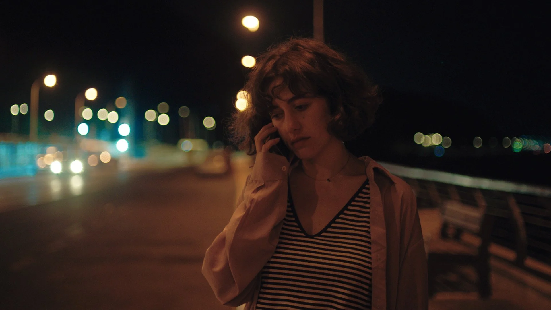 Une jeune femme avec des cheveux bouclés bruns parle au téléphone la nuit sur un quai, avec des lampadaires et des lumières floues en arrière-plan.