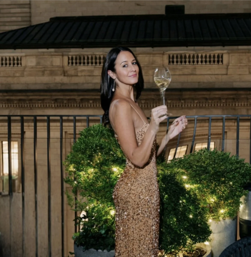Woman in a gold sequin dress holding a glass of white wine on a balcony with decorated green plants and a city building in the background.