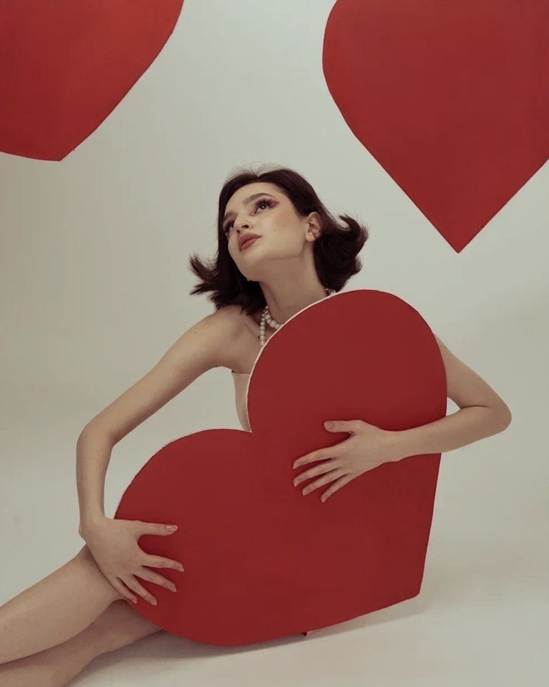 A woman with dark hair and makeup, wearing a pearl necklace, holding a large red heart-shaped object, posed with a white background and red heart cutouts.