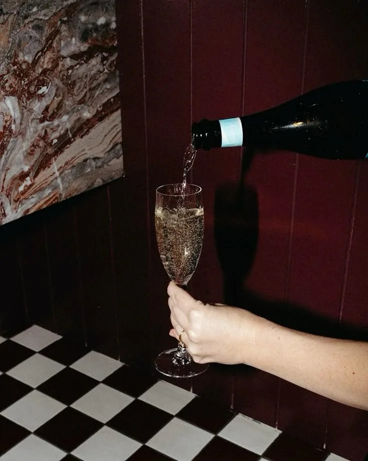 Person holding a champagne flute below a bottle of champagne being poured into it, with a dark red wooden wall and black-and-white checkered floor in the background.