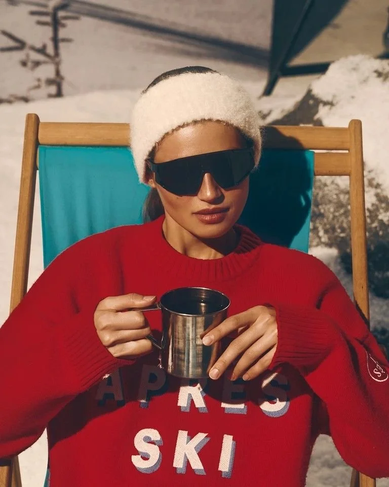 Woman wearing a white headband and black sunglasses, sitting outdoors on a blue deck chair, holding a metallic mug, wearing a red sweater with the words 'APRES SKI' in large letters, snow visible in the background.
