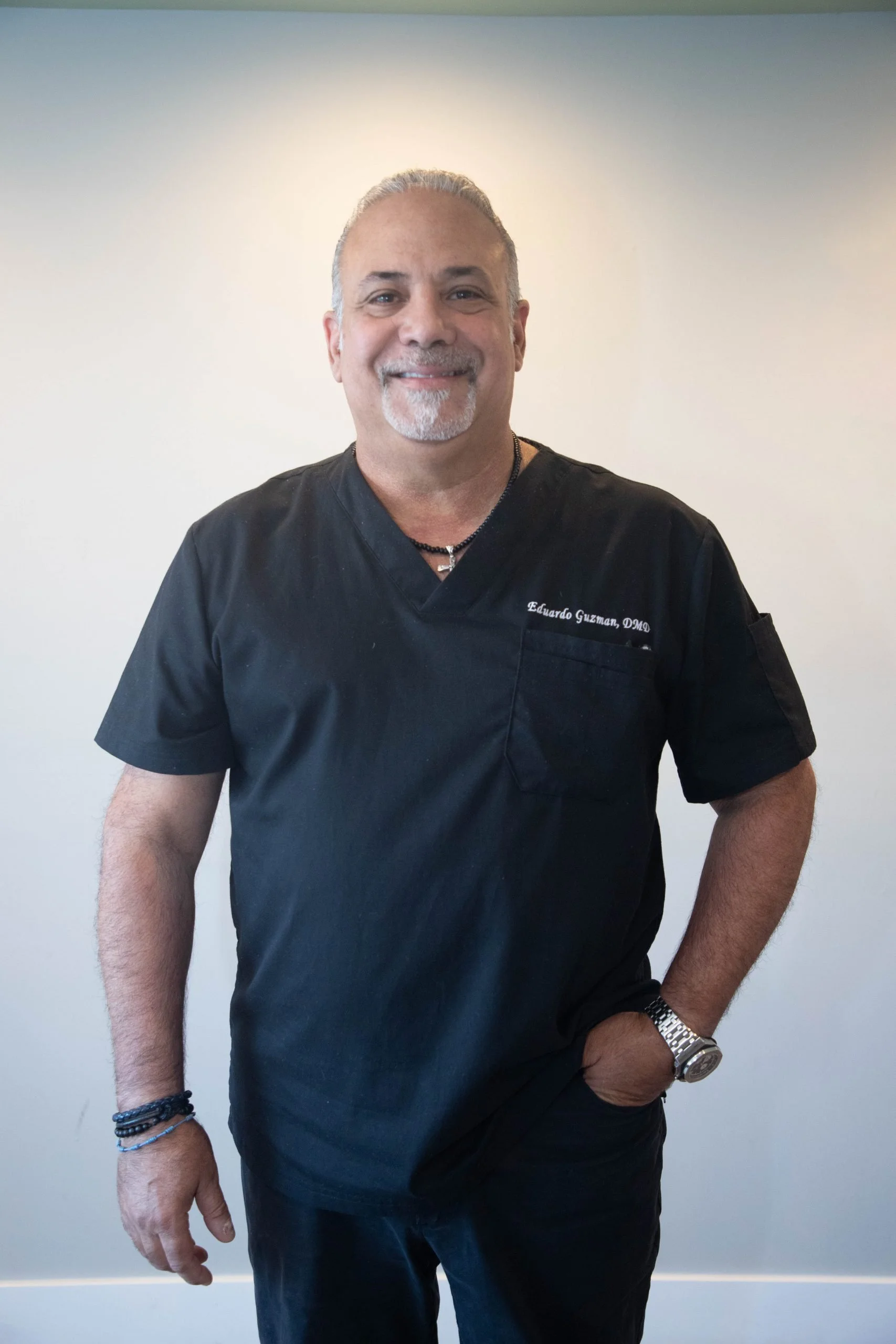 A smiling man in black scrubs standing against a plain wall, wearing a wristwatch and bracelets, with 'Eduardo Guzman, DMD' embroidered on his scrubs.