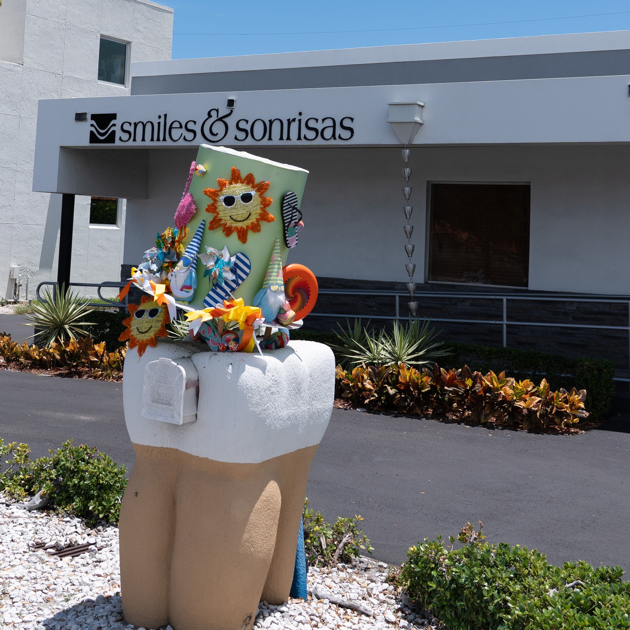 Decorative oversized tooth sculpture outside a dental office, decorated with colorful party items and smiling sun faces, in front of a building with a sign that reads 'smiles & sonrisas'.