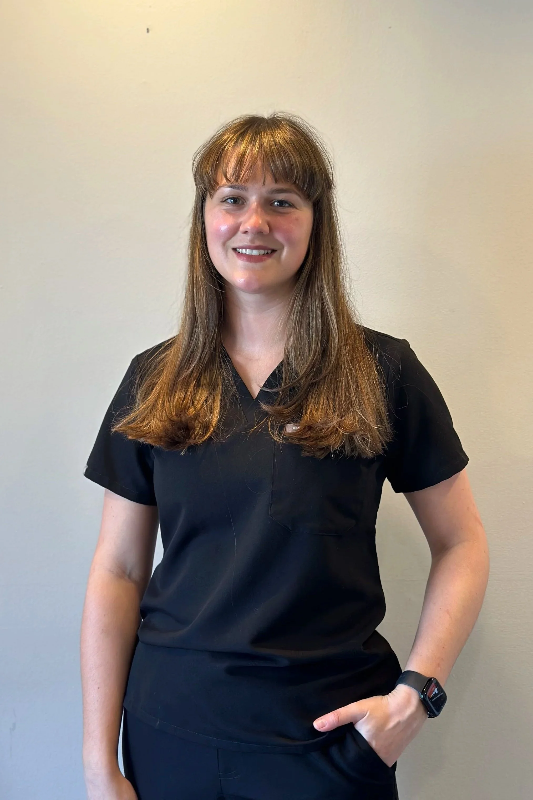 A young woman with brown hair and bangs wearing a black casual uniform top posing against a plain off-white wall.