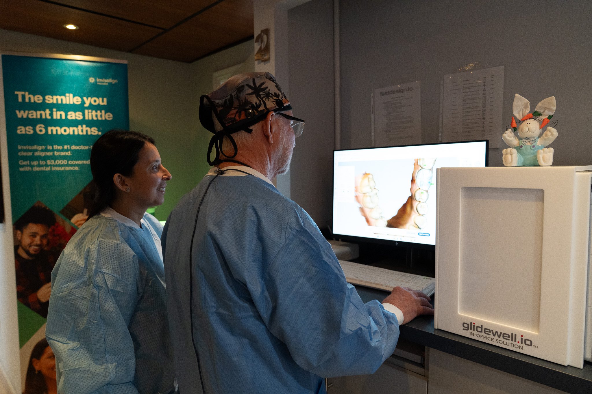 Two dental professionals, a woman and an older man, examine a digital dental scan on a computer monitor in a dental clinic. The woman has dark hair and is smiling, wearing a blue gown. The man is wearing a patterned surgical cap, glasses, and a blue gown. A promotional poster about Invisalign is visible in the background.