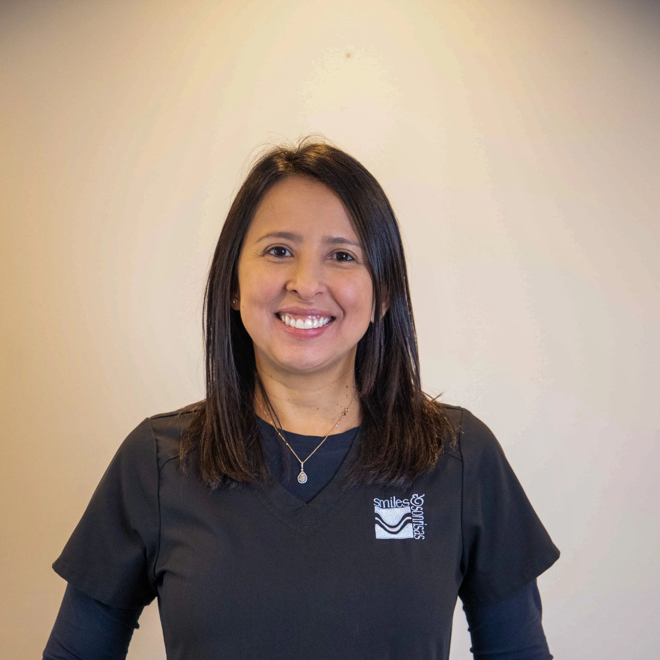 A woman with shoulder-length dark hair smiling at the camera, wearing a black scrub top with a dental logo, standing against a plain light-colored wall.