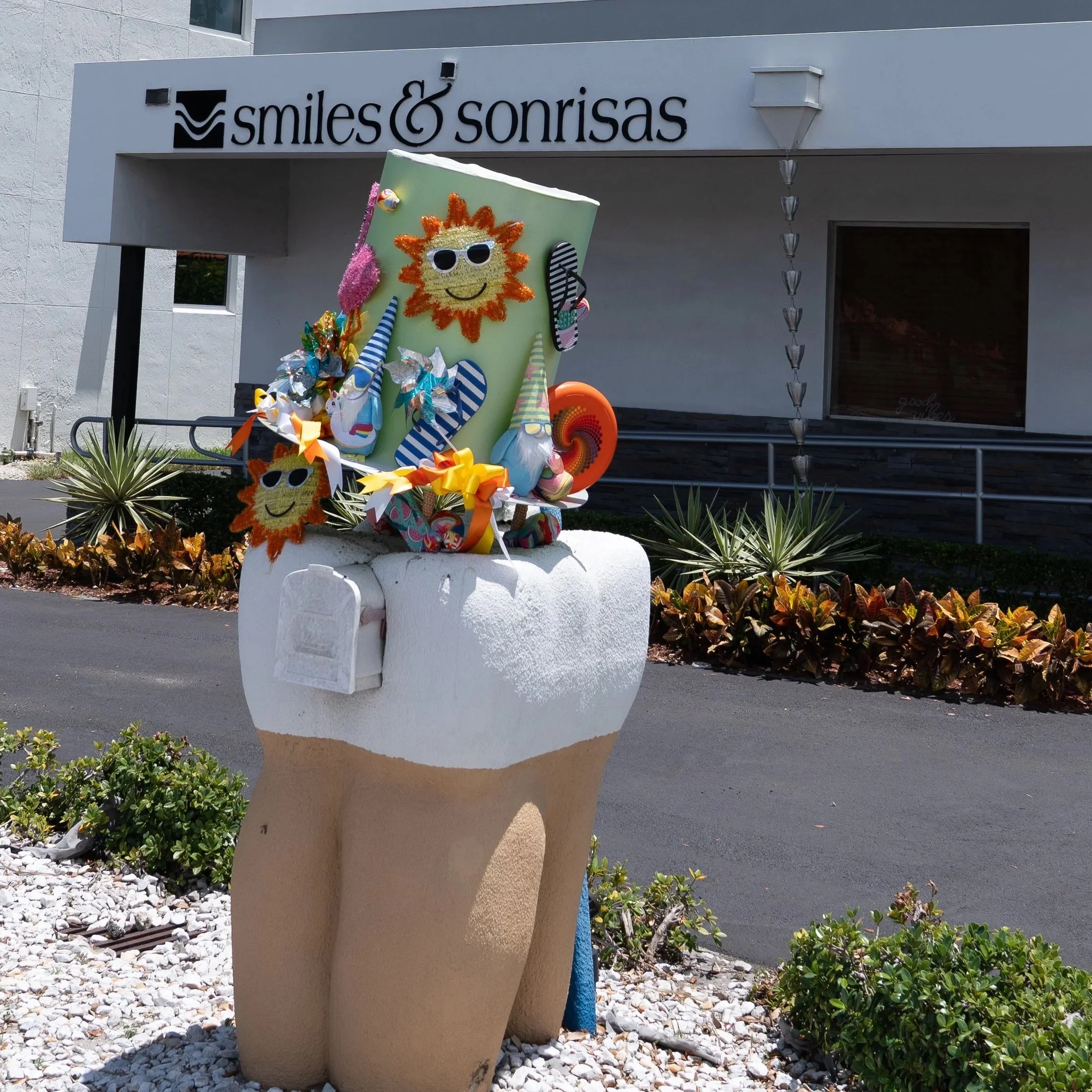 Decorative figurine of a large tooth with a smiling face, sunglasses, and a sun design on the top, placed outdoors with plants and a building in the background. The figurine is colorful and whimsical.