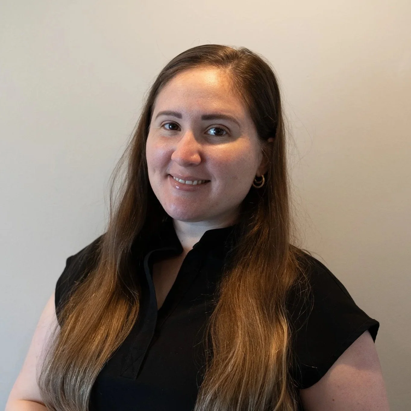 A woman with long brown hair, wearing a black scrub top and gold jewelry, smiling in front of a textured blue and gray backdrop.