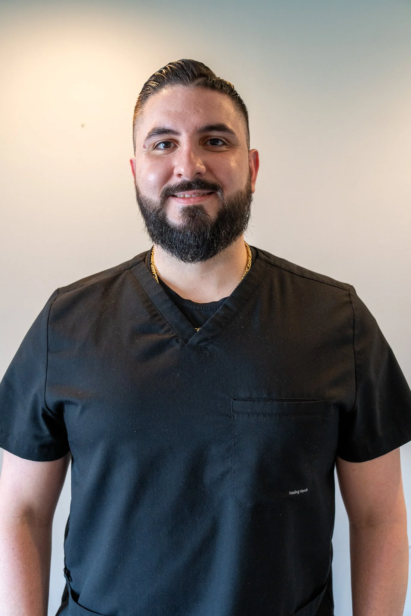 A young woman with long dark hair and a bright smile, wearing a black top, standing against a plain white wall.