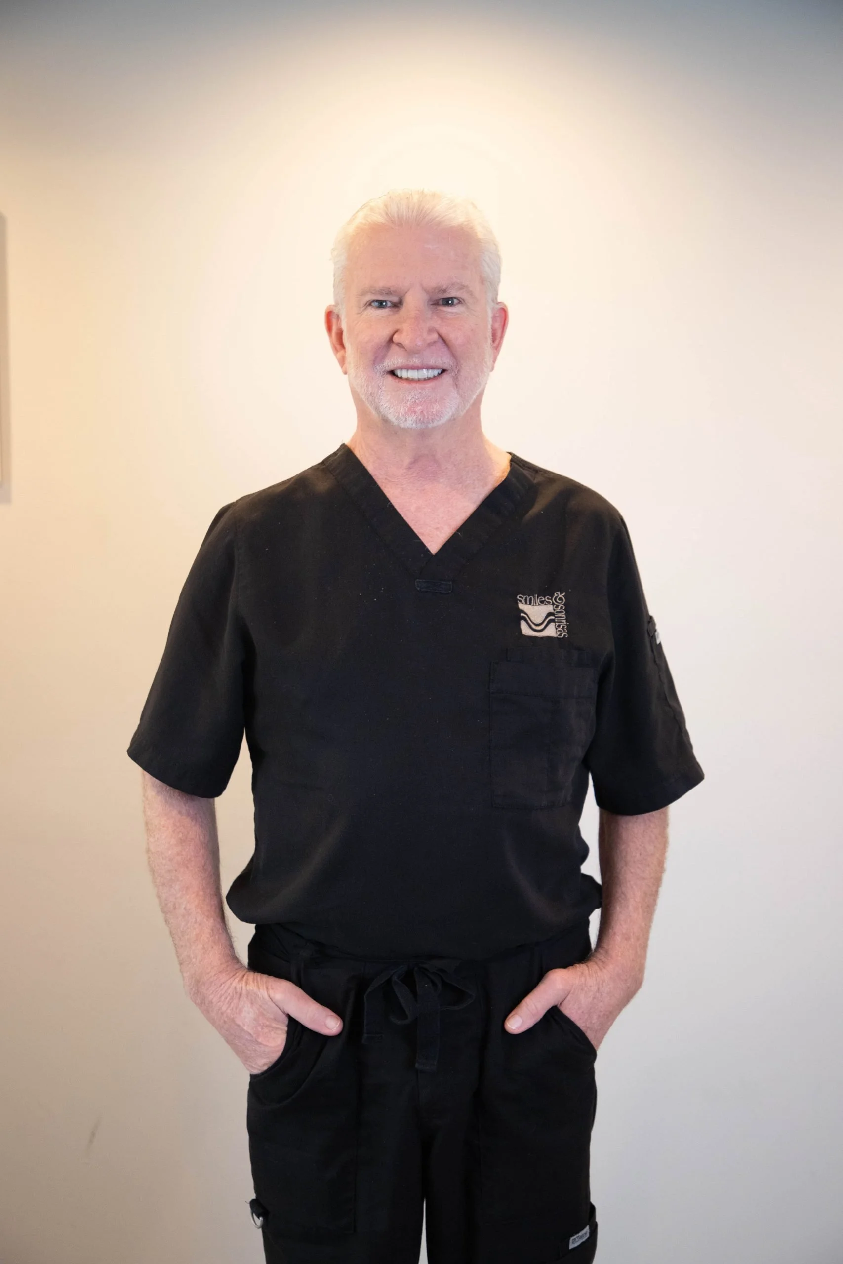 A smiling elderly man with white hair and beard wearing black medical scrubs standing against a plain light-colored wall.