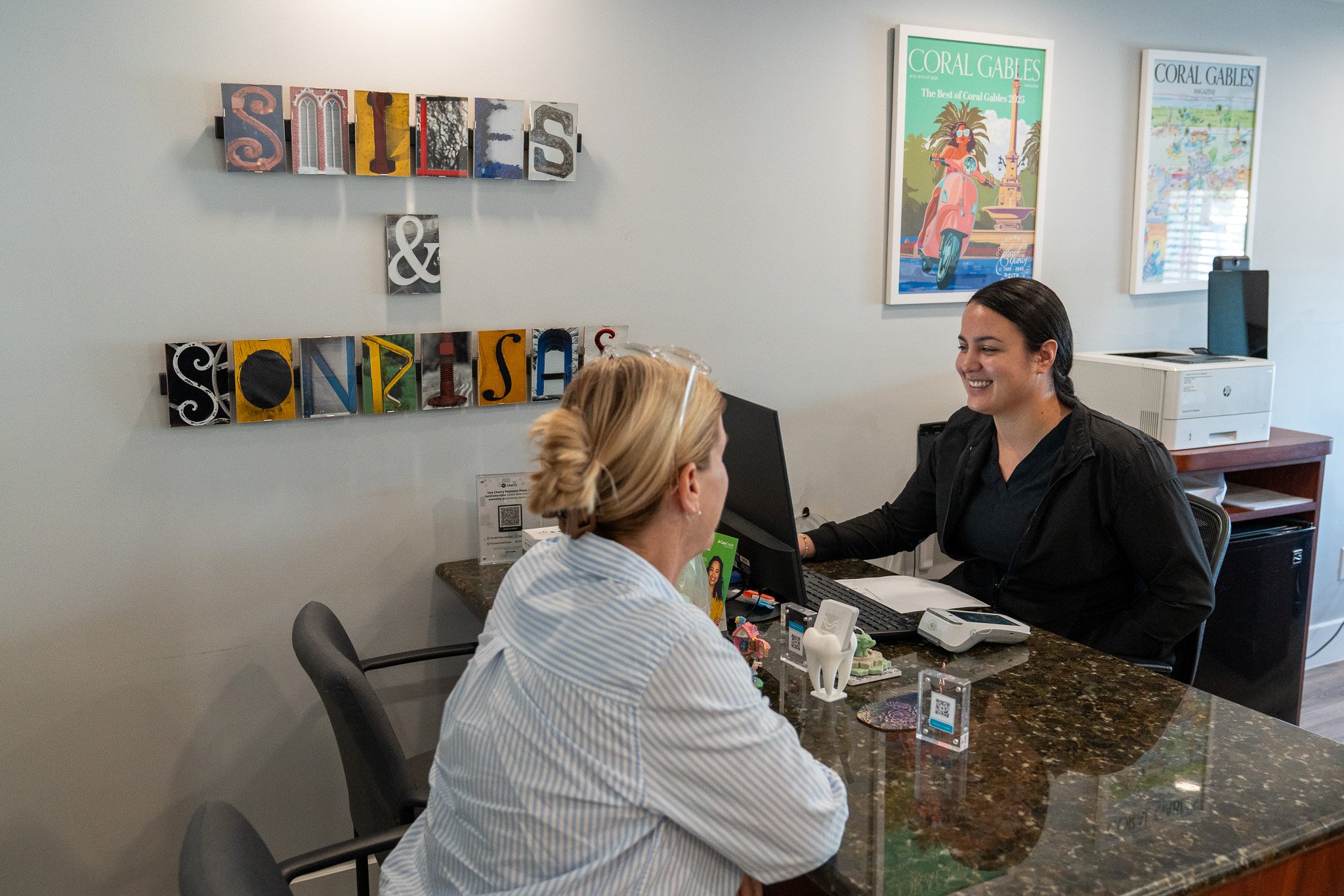 Two women sitting at a reception desk having a conversation, with decorative wall art and colorful posters in the background.