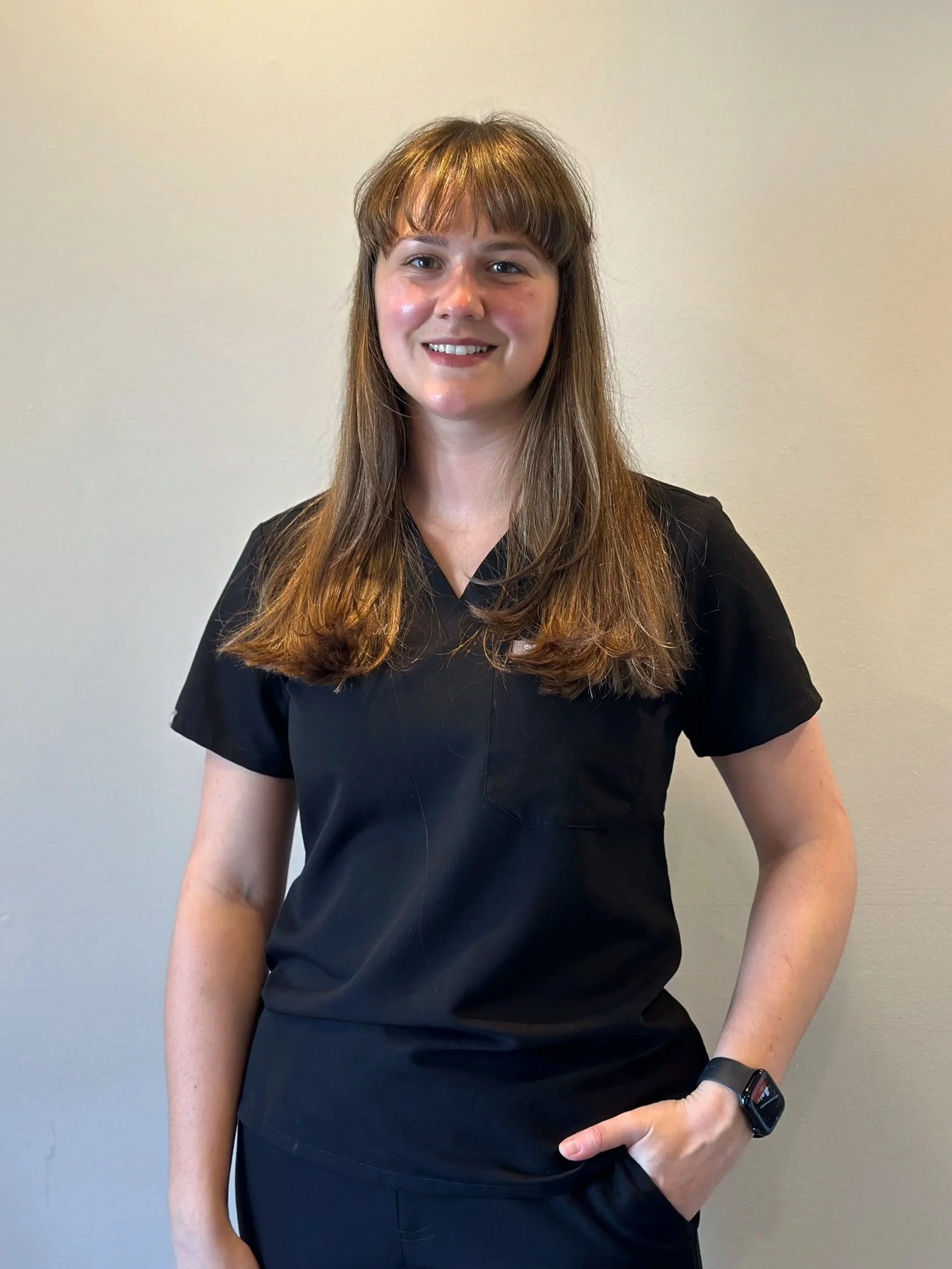 A woman with long brown hair, wearing a black medical scrub top and smartwatch, smiling and standing against a plain light-colored wall.
