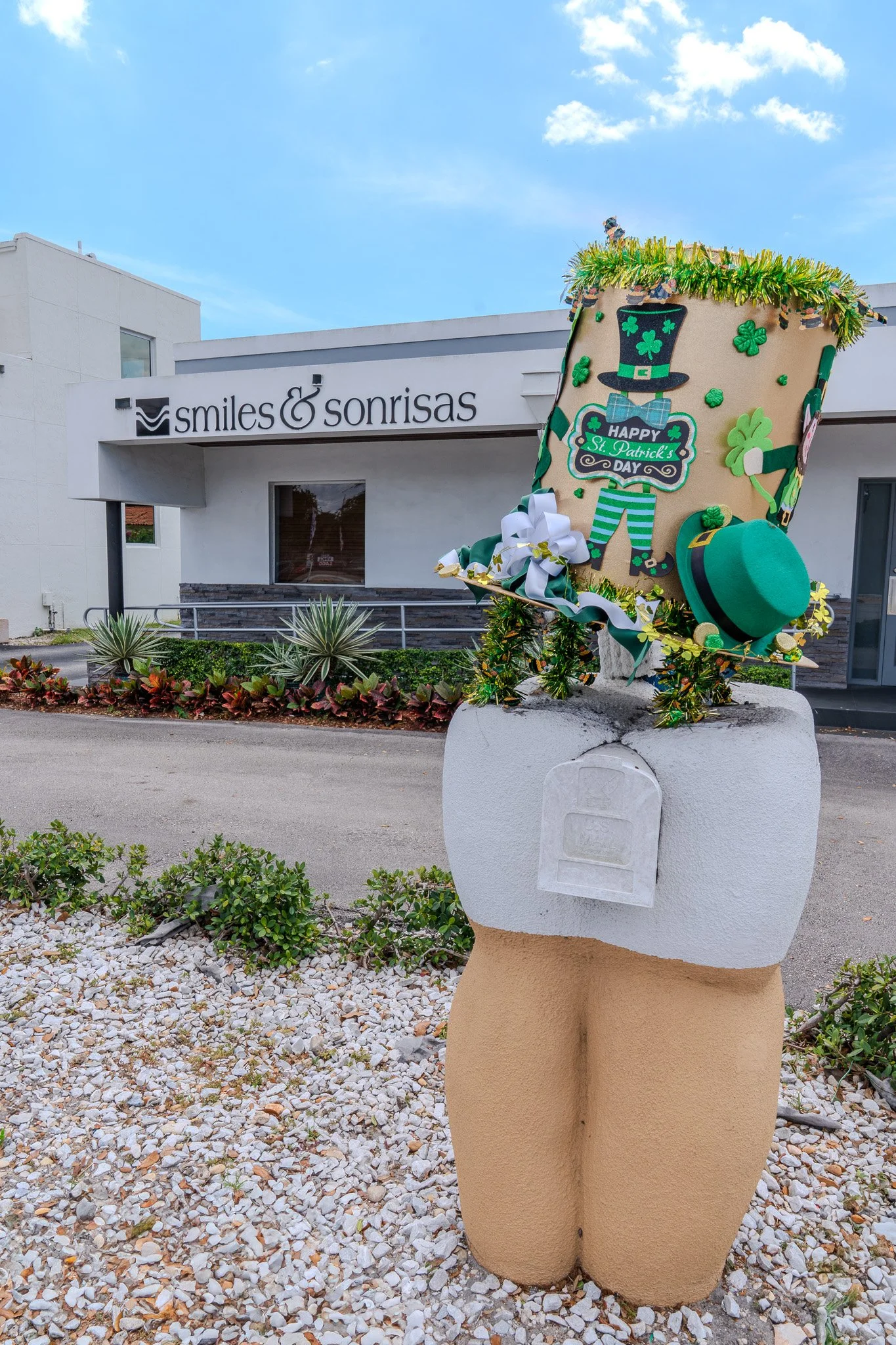 Decorative St. Patrick's Day urn with leprechaun, hat, shamrocks, and a 'Happy St. Patrick's Day' sign in front of a dental office sign that reads 'smiles & sonrisas.'