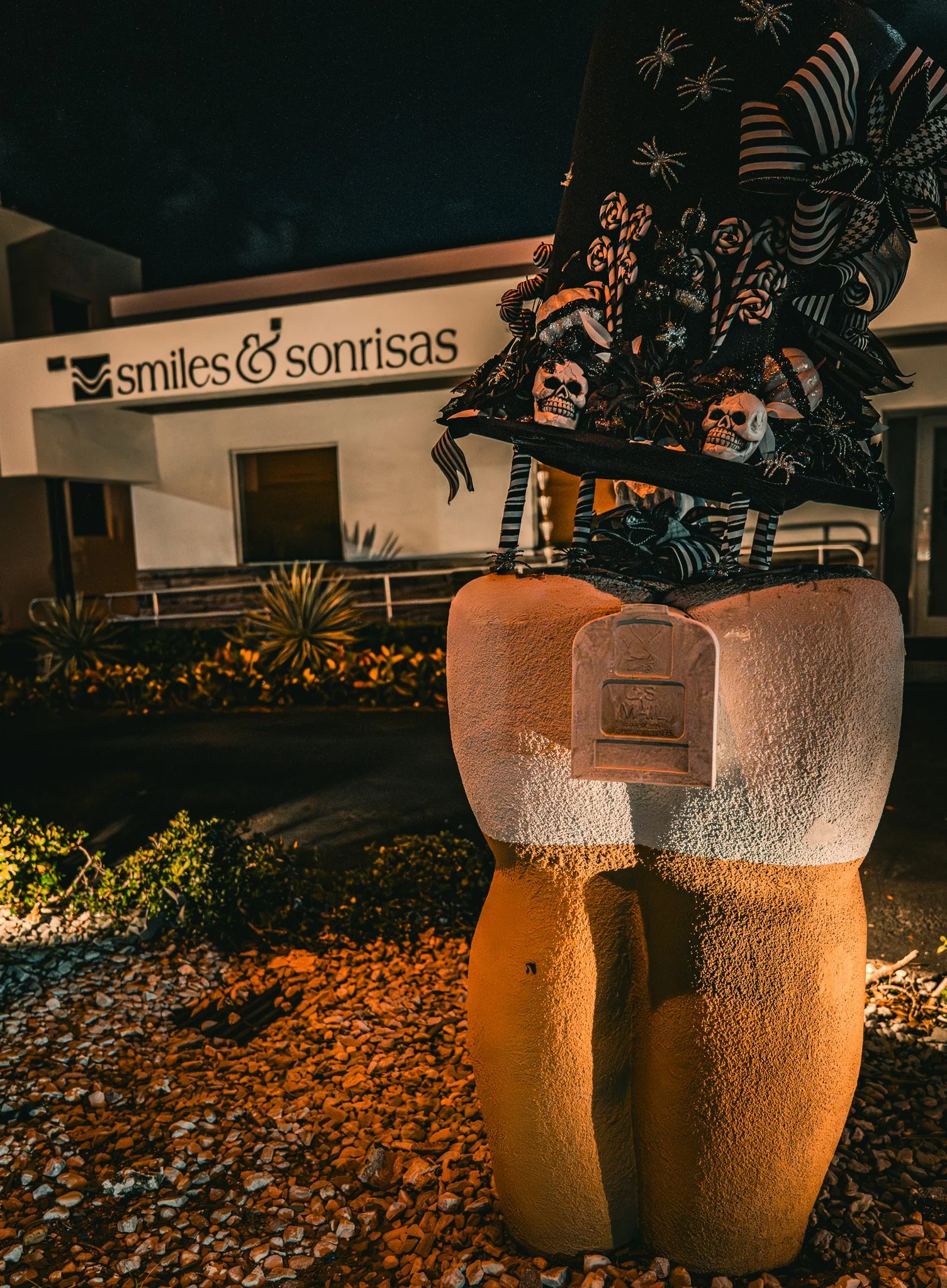 A Halloween-themed sculpture of a tooth with a witch's hat made of striped ribbons, candy canes, skulls, and black flowers, situated outside a dental office named 'smiles & sonrisas' in the evening.