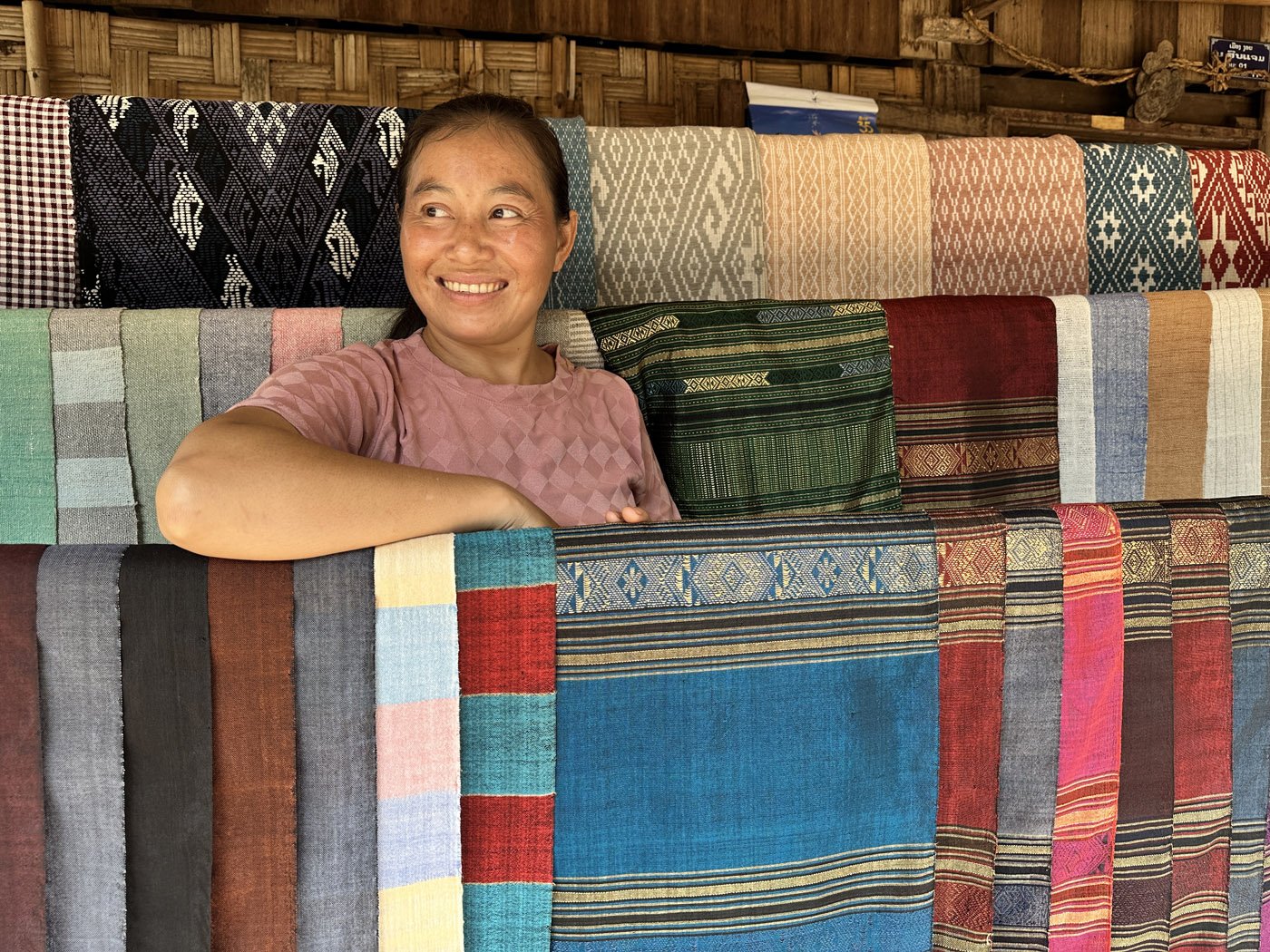 A smiling woman leaning on a display of colourful handloom woven fabrics in a market in Laos.