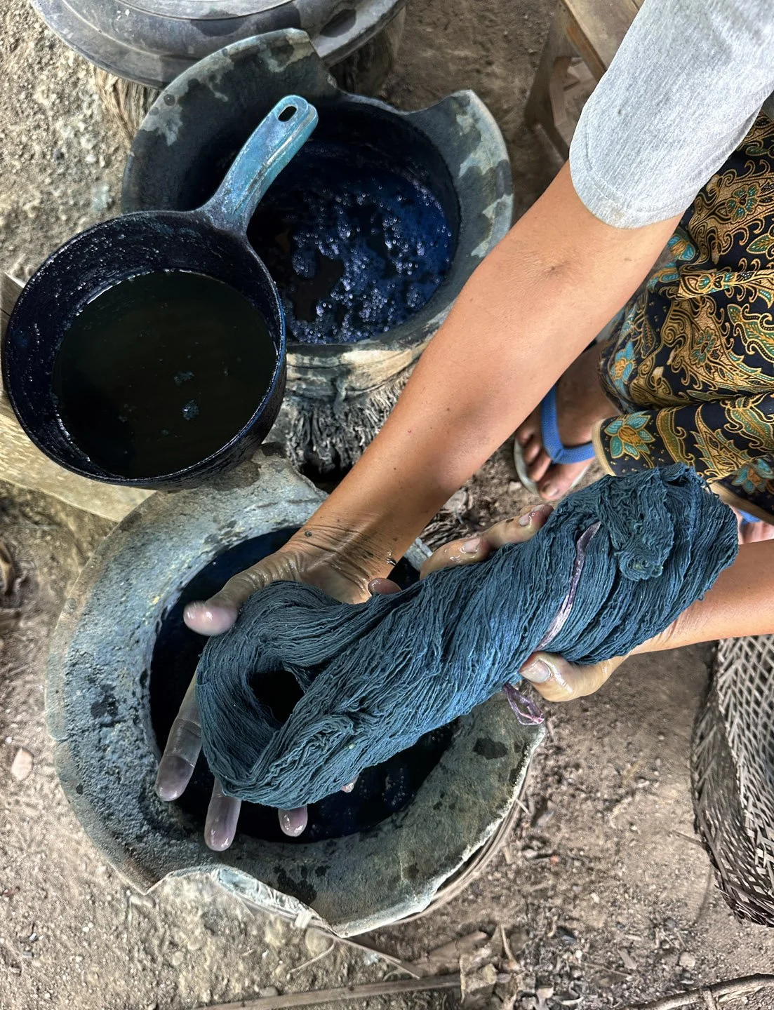 Hand dyeing cotton in organic indigo blue dye outdoors for traditional cloth weaving, with pots of natural dye nearby.