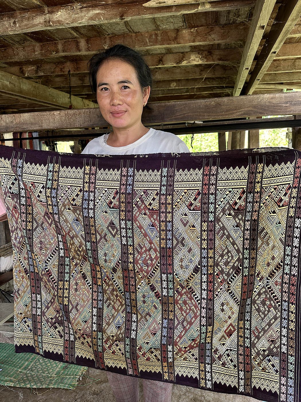 Woman holding a large silk, colourful, intricately patterned, handloom woven textile in a rustic workshop.