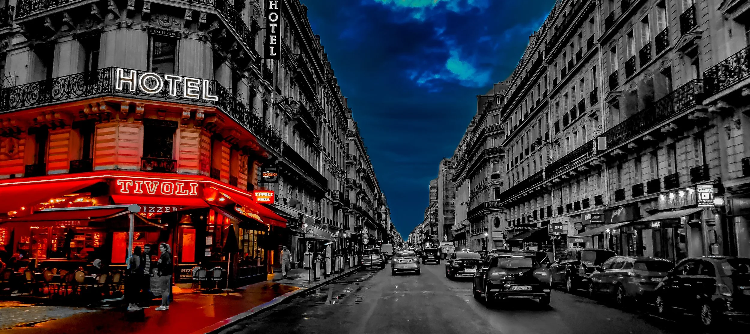 City street with multiple parked cars, tall buildings on both sides, a hotel sign, and a restaurant with neon lights, under a cloudy blue sky at dusk.