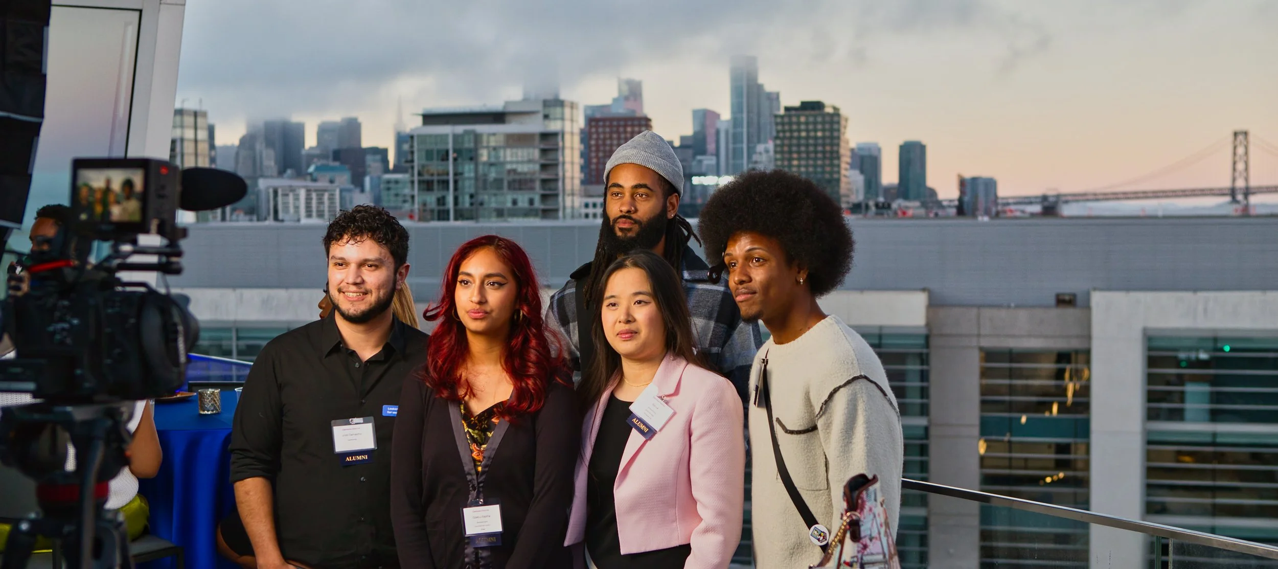 A group of six diverse young adults standing together on a rooftop with a city skyline in the background, during what appears to be a professional or alumni event.