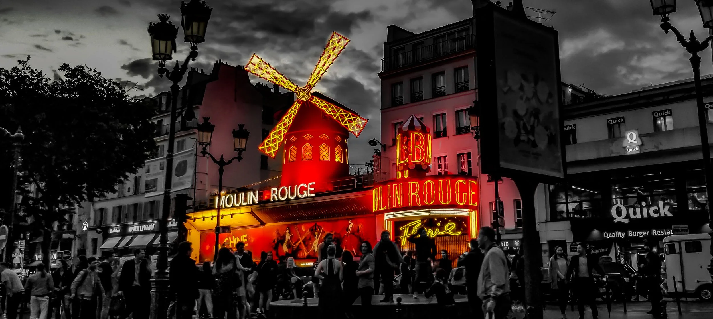 Night view of the Moulin Rouge in Paris with neon signs and a brightly lit windmill on top. The image is mostly in black and white, with the neon signs and windmill illuminated in red and yellow. There are people walking and gathering in front of the